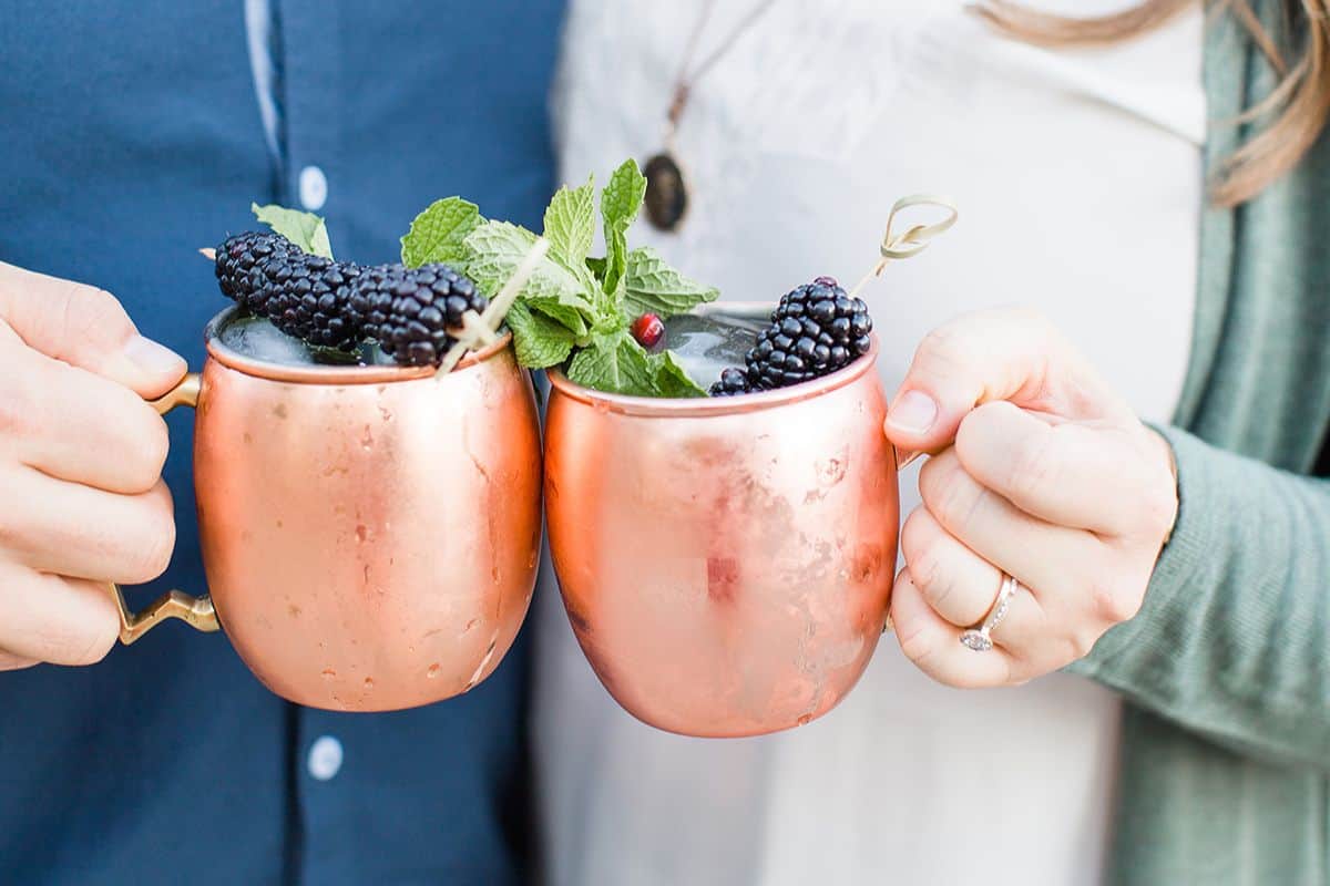 A close up of a man and woman's hand each holding a copper Moscow Mule mug with blackberry and mint garnish.