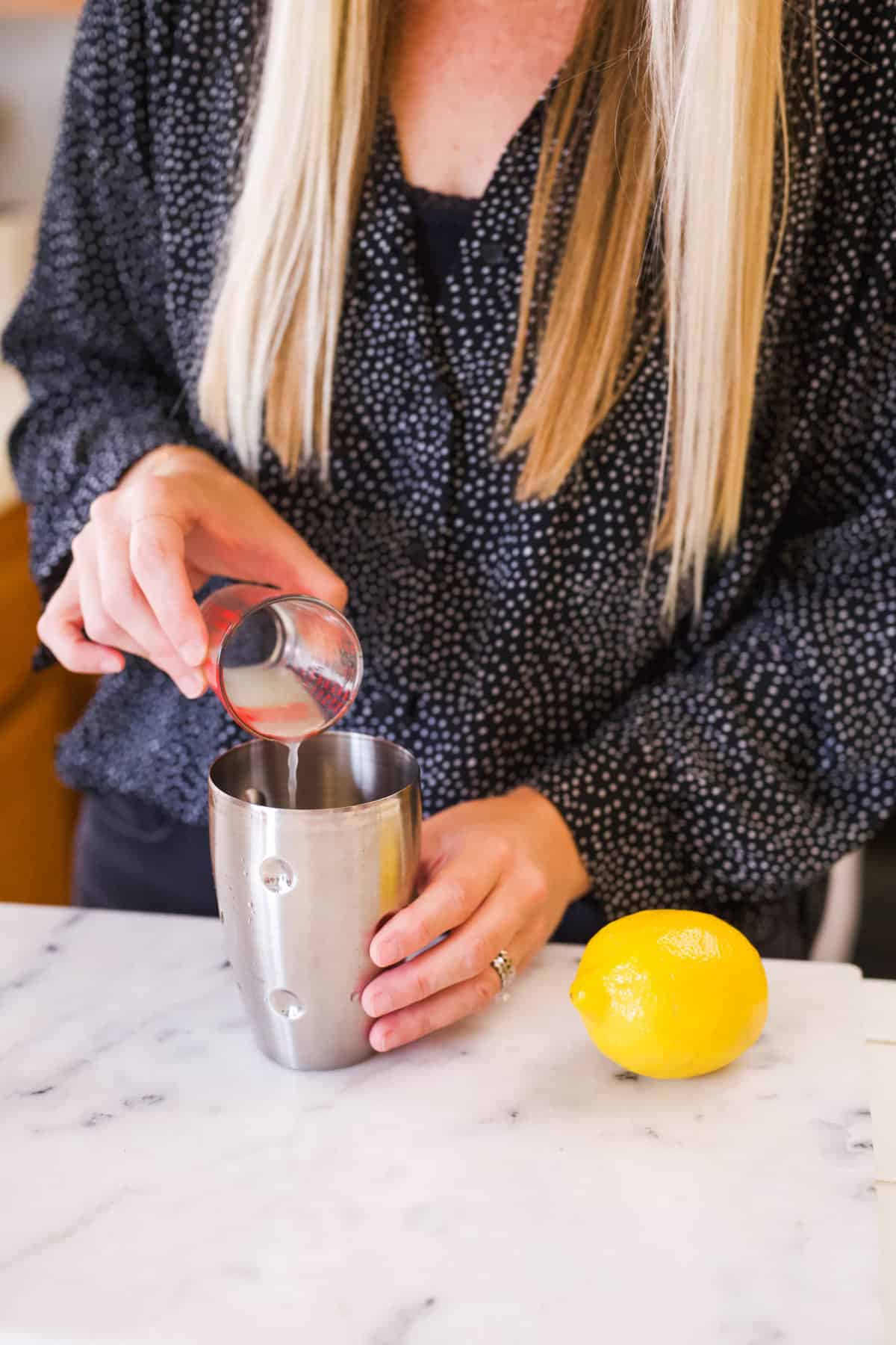 Woman adding fresh lemon juice to a cocktail shaker from a jigger at a counter.