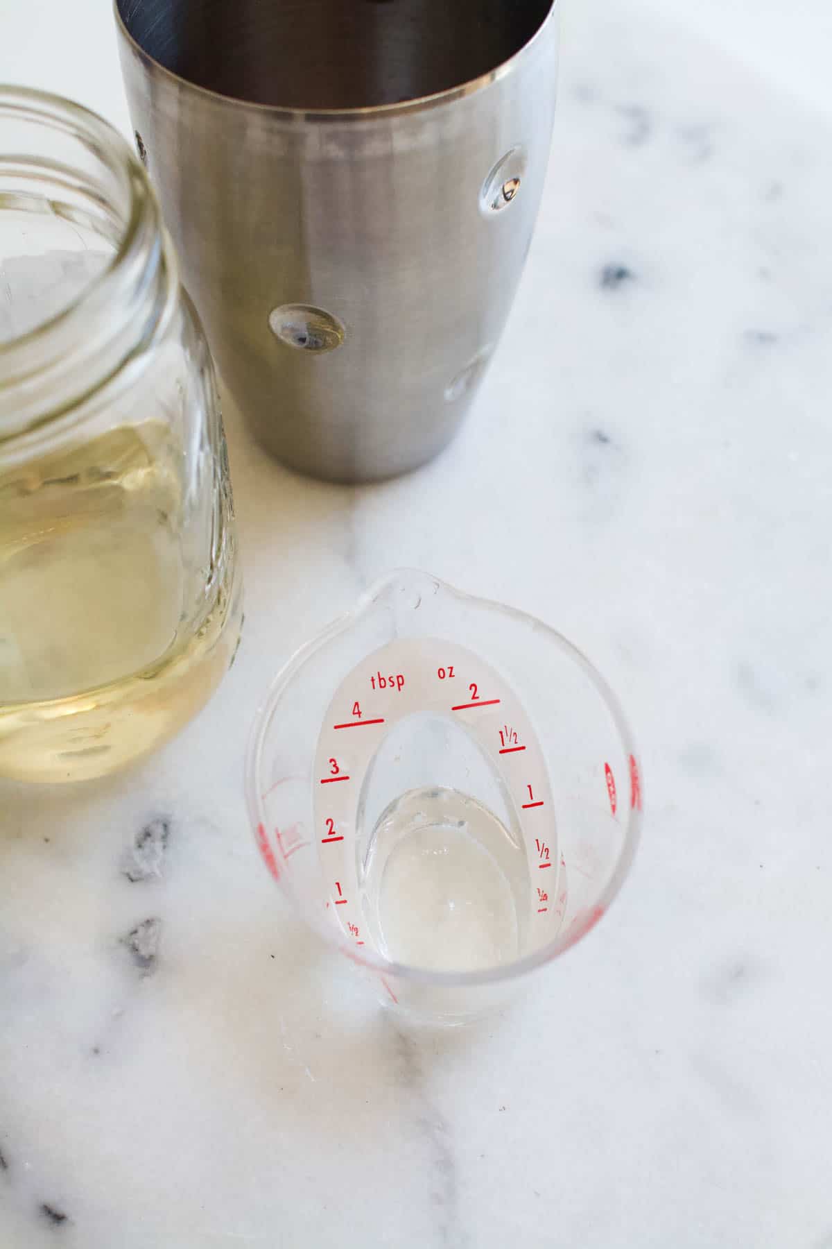 A measuring cup with simple syrup on a counter next to a jar of syrup and a cocktail shaker.