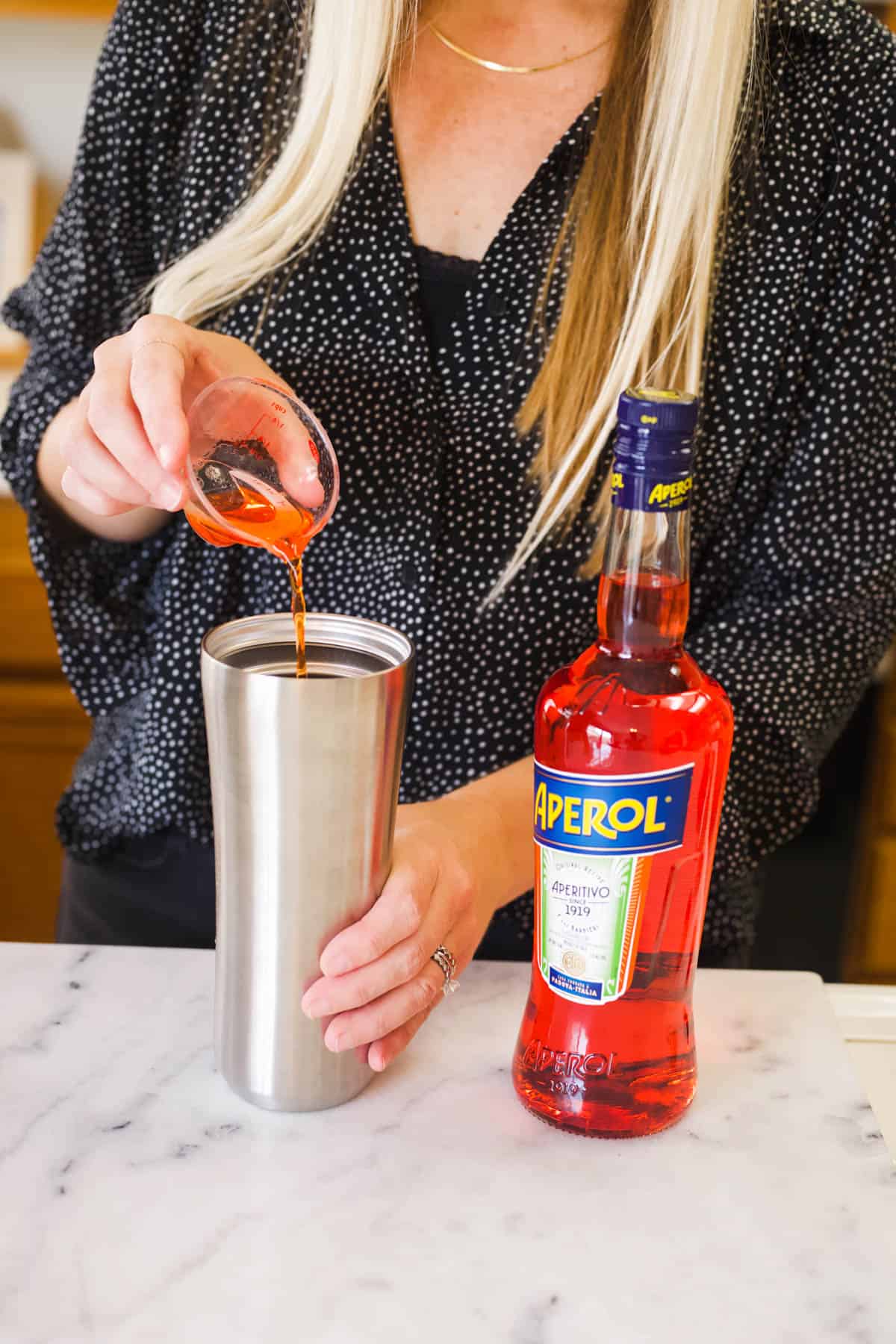 A woman adding Aperol to a cocktail shaker next to a large bottle of Aperol liqueur.