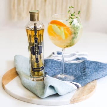 A Cointreau spritz recipe in a large wine glass and next to a bottle of St Germain on top of a blue towel and wood and marble serving tray.