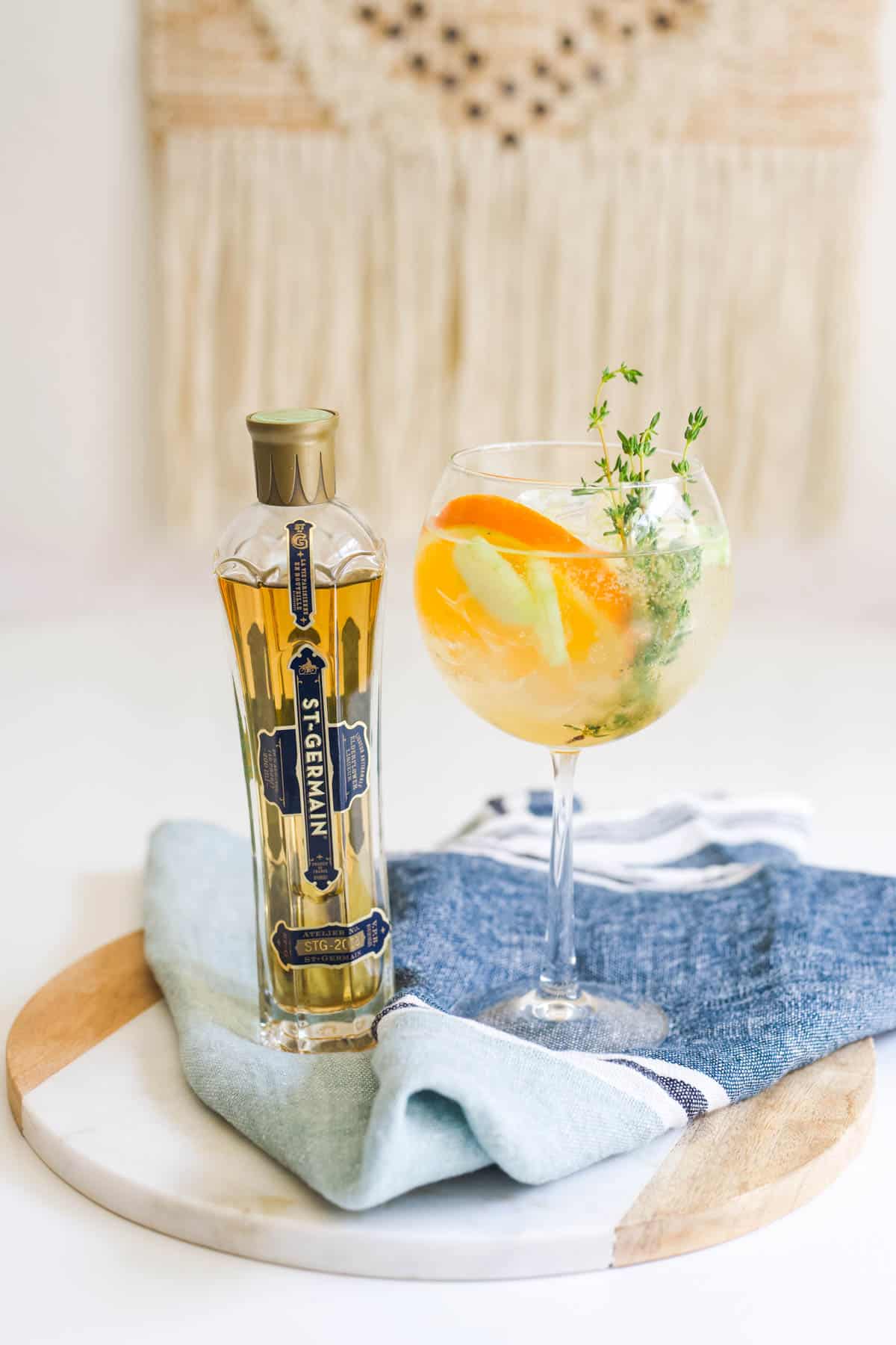 A Cointreau cocktail with cucumber and St Germain next to a bottle of St Germain, all on top of a marble and wood serving board with a blue towel.