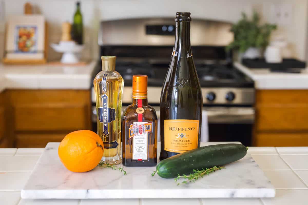 Ingredients you will need to make a Cointreau cocktail including a fresh orange, a bottle of St Germain elderflower liqueur, a bottle of Cointreau, a bottle of prosecco, and fresh cucumber.