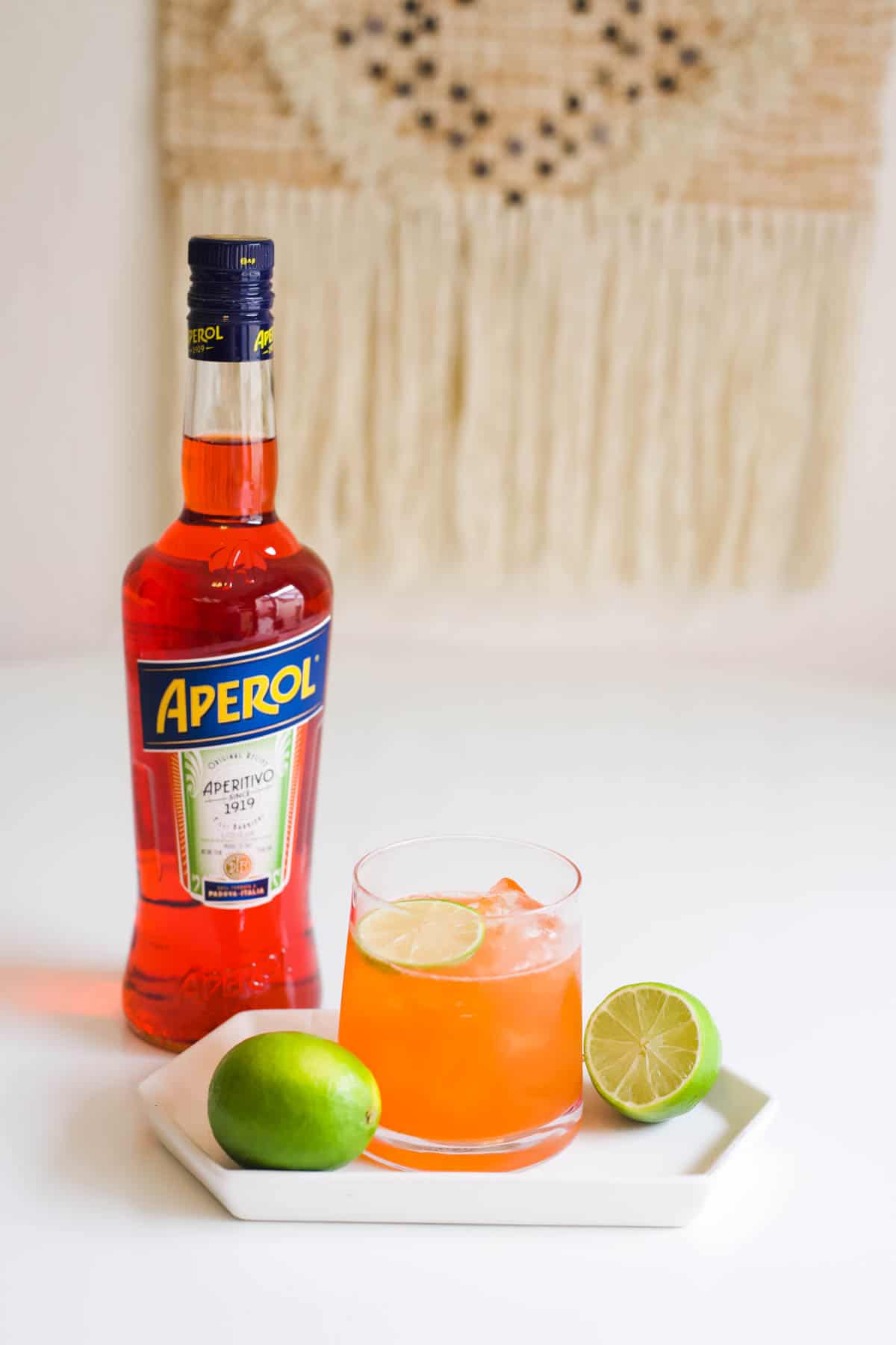 A small white plate on a white counter top topped with a glass of a rum Aperol cocktail, fresh limes and topped with a lime slice in front of a bottle of Aperol liqueur.