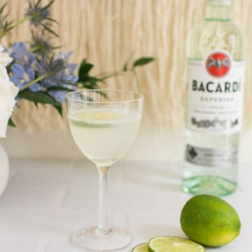 Classic Daiquiri in a gimlet glass next to a sliced lime and a bottle of white rum.