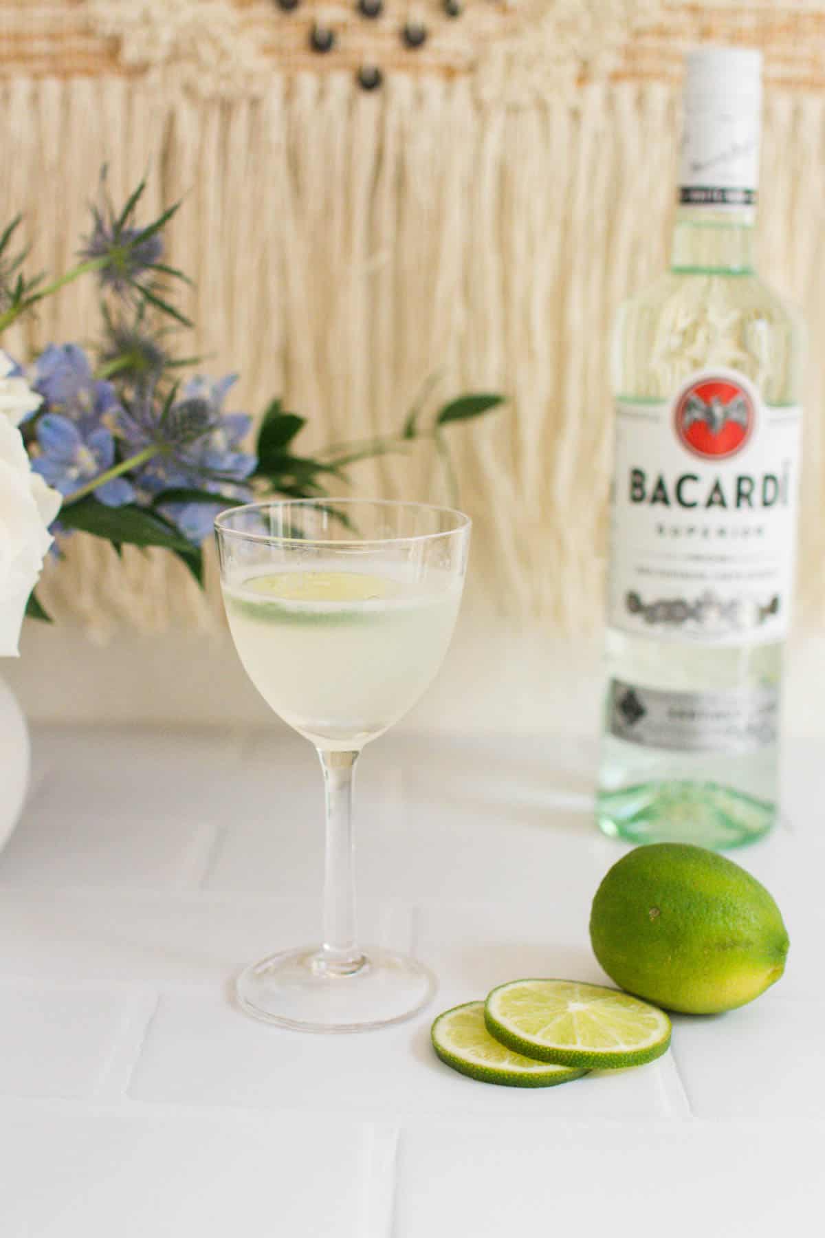 Classic Daiquiri in a gimlet glass next to a sliced lime and a bottle of white rum.