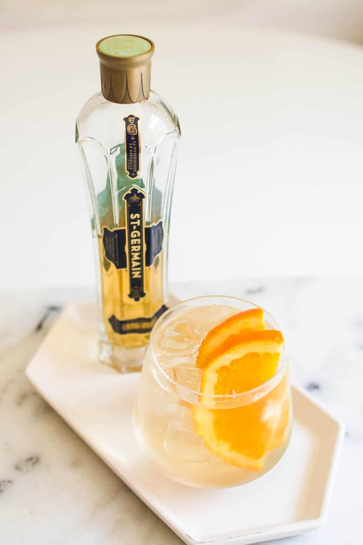 A glass of White Wine St. Germain spritzer garnished with orange slices in front of a bottle of St. Germain liqueur.