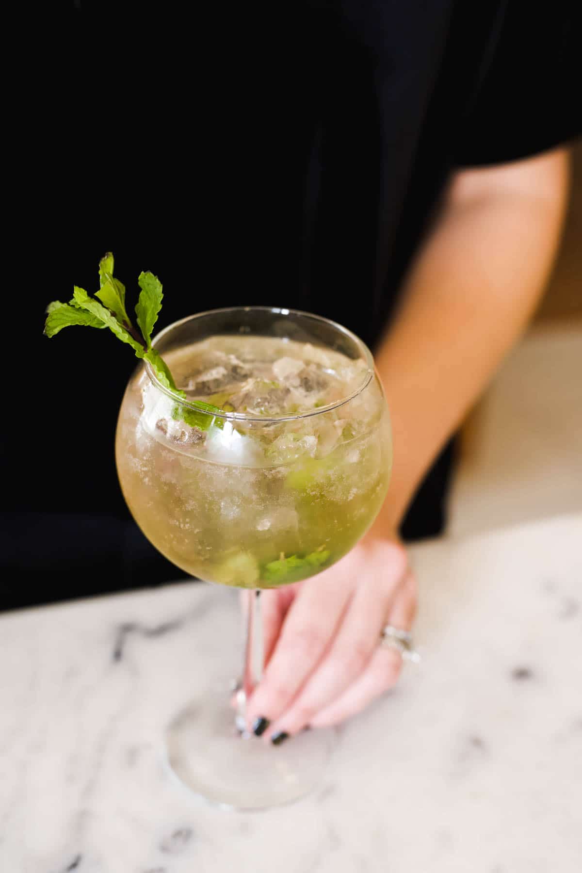 Woman pushing a Hugo Spritz garnished with fresh mint across the counter towards you.