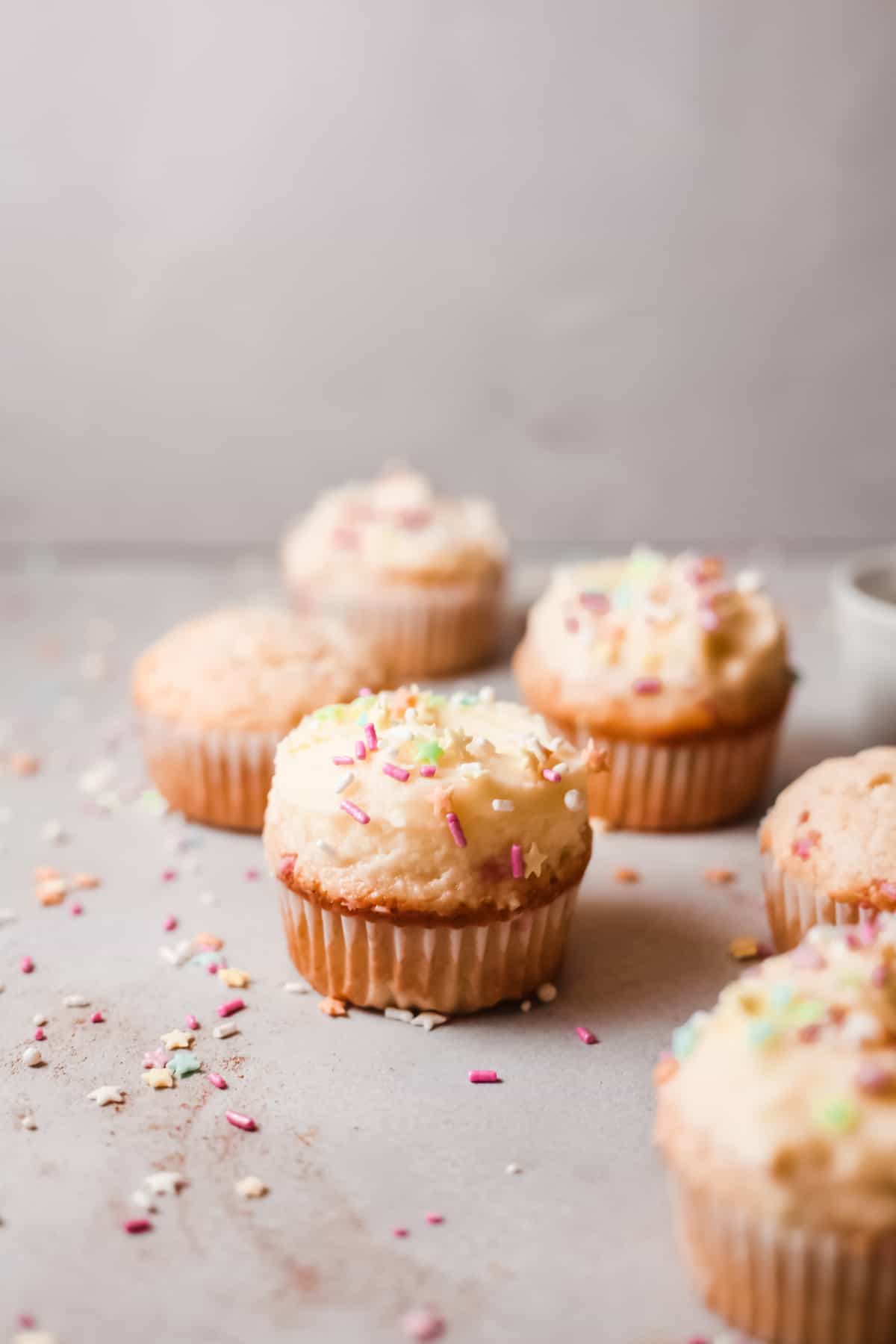 Six birthday cupcakes with sprinkles on a gray countertop surface.