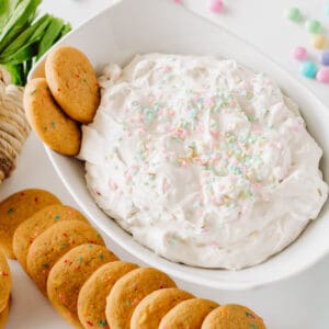 A white bowl holding a cake mix and cream cheese dessert dip for Easter with cookies around it.