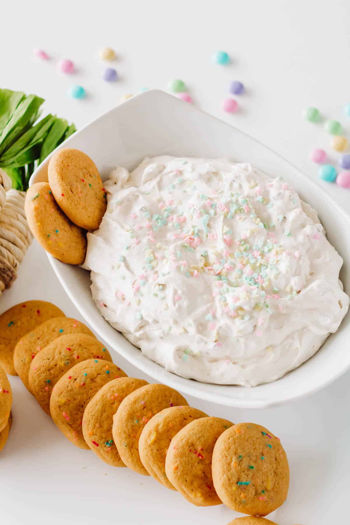 A white bowl with an Easter dessert dip topped with sprinkles on a table next to cookies.