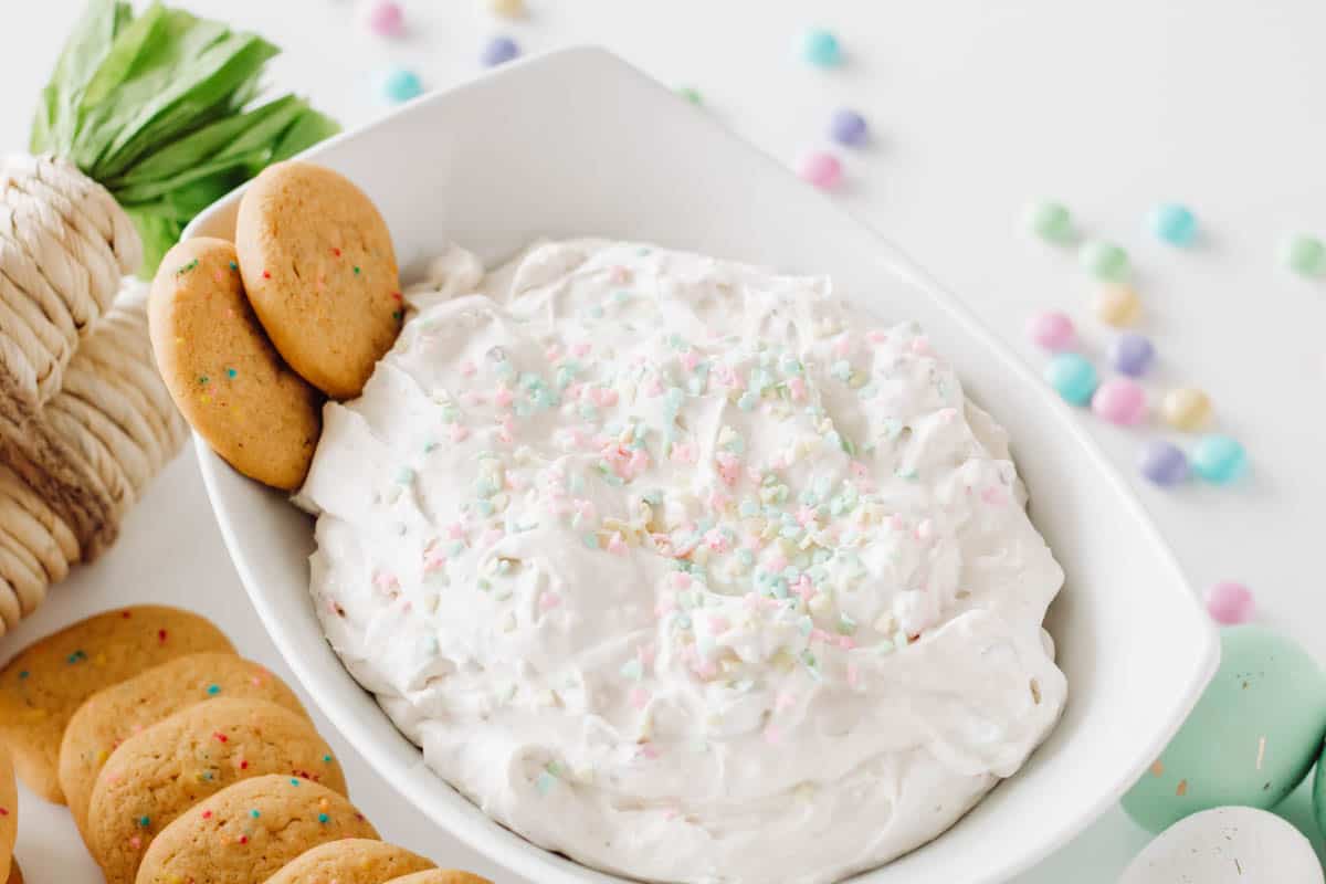 A cream cheese dip topped with Easter sprinkles in a white bowl on a table with cookies around it and 2 stuck in the dip.