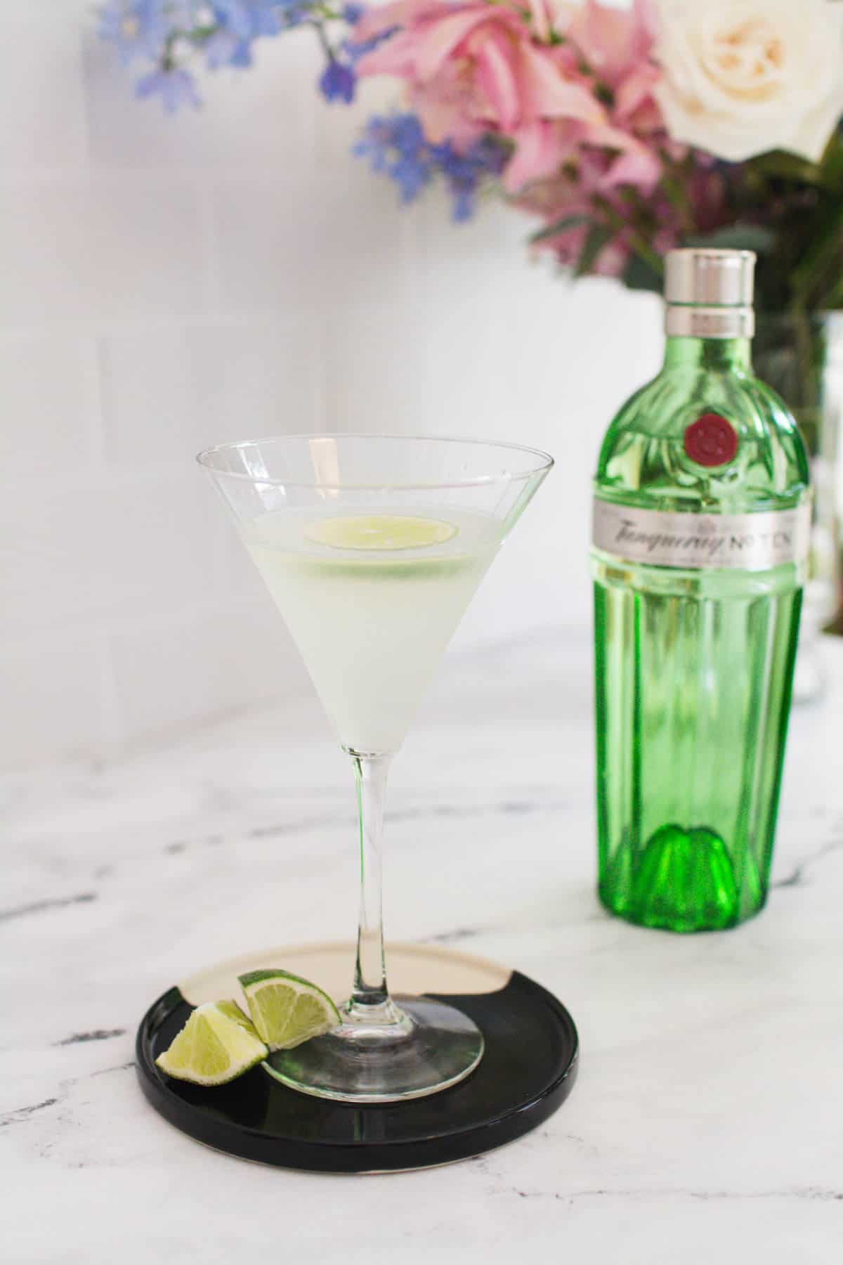 Gin gimlet in a martini glass with a bottle of gin and bunch of flowers in the background.