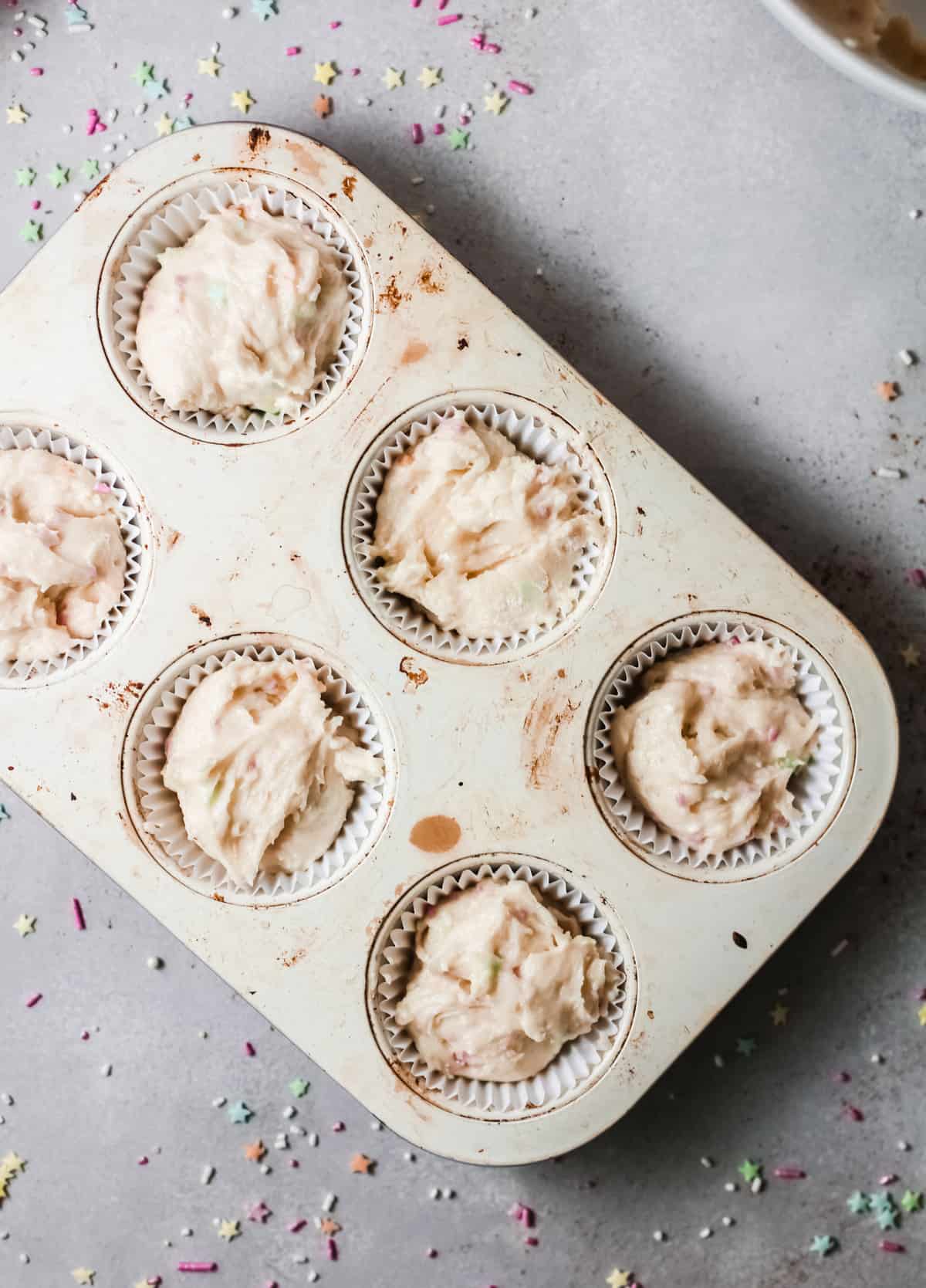 A 6-well muffin tin filled with paper liners and birthday cake batter with sprinkles ready for baking.