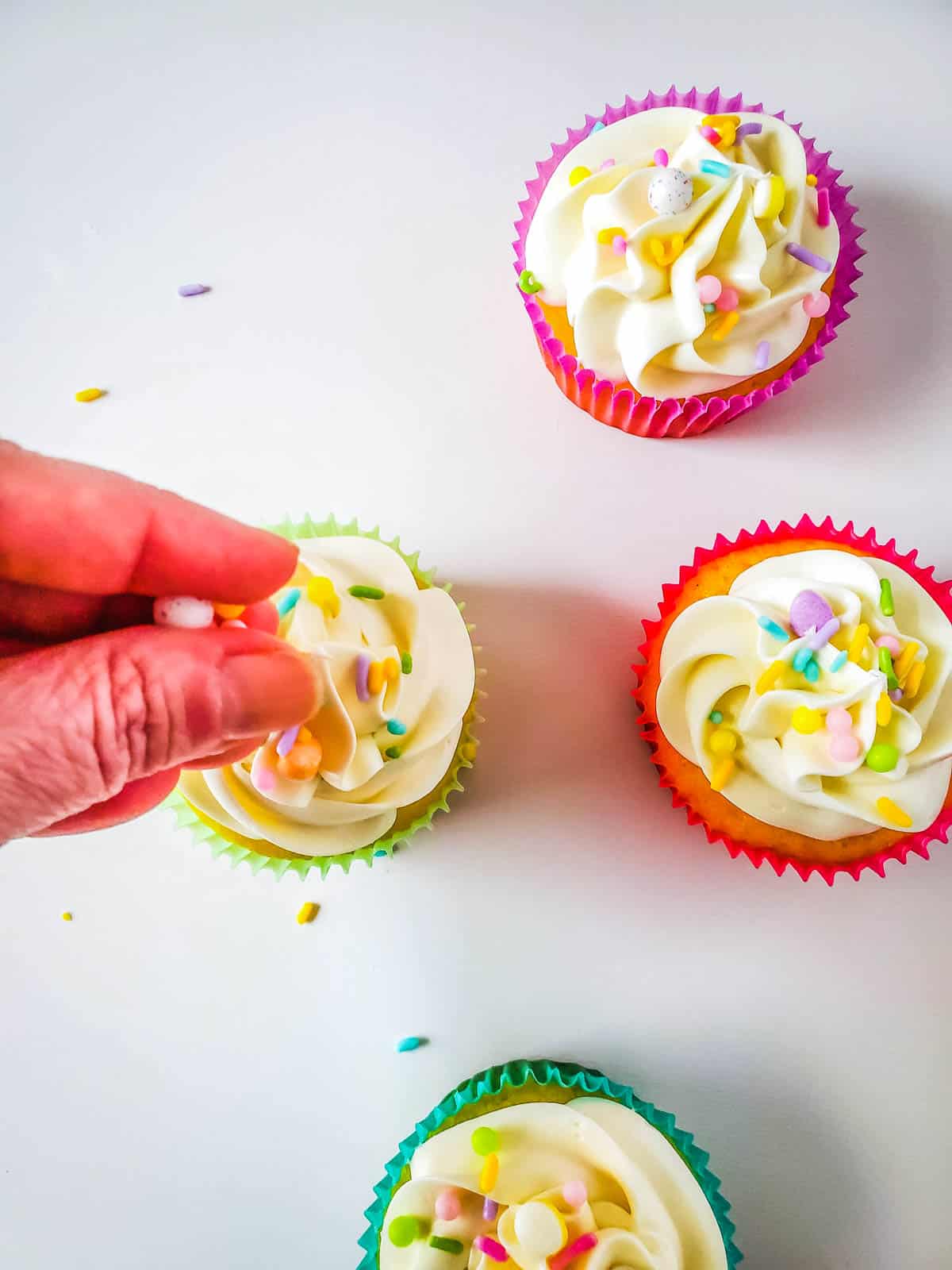 Frosted cupcakes on a table with a woman adding Easter sprinkles to decorate.