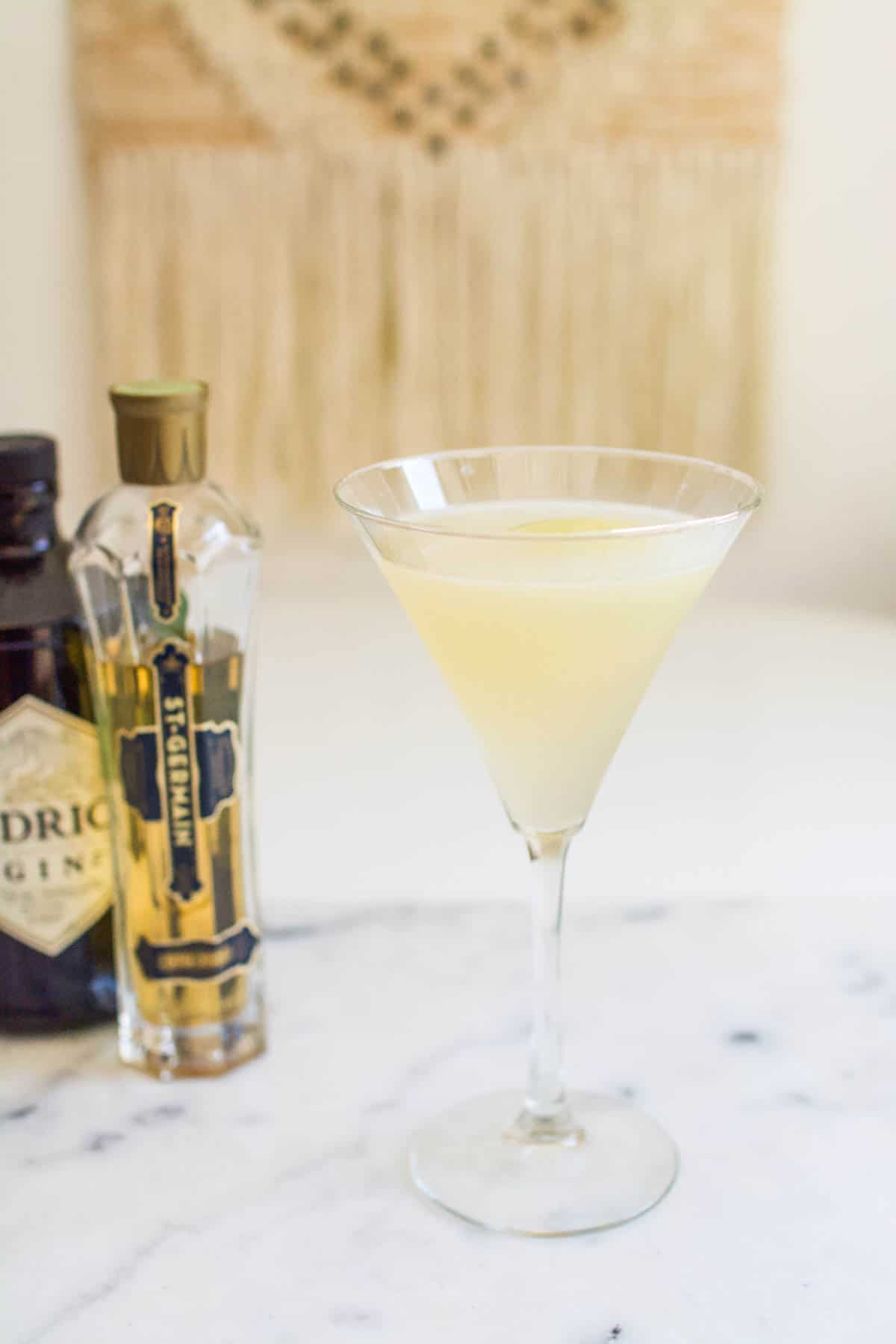 A St. Germain French gimlet cocktail in a martini glass on a table with a bottle of St Germain and gin behind it.