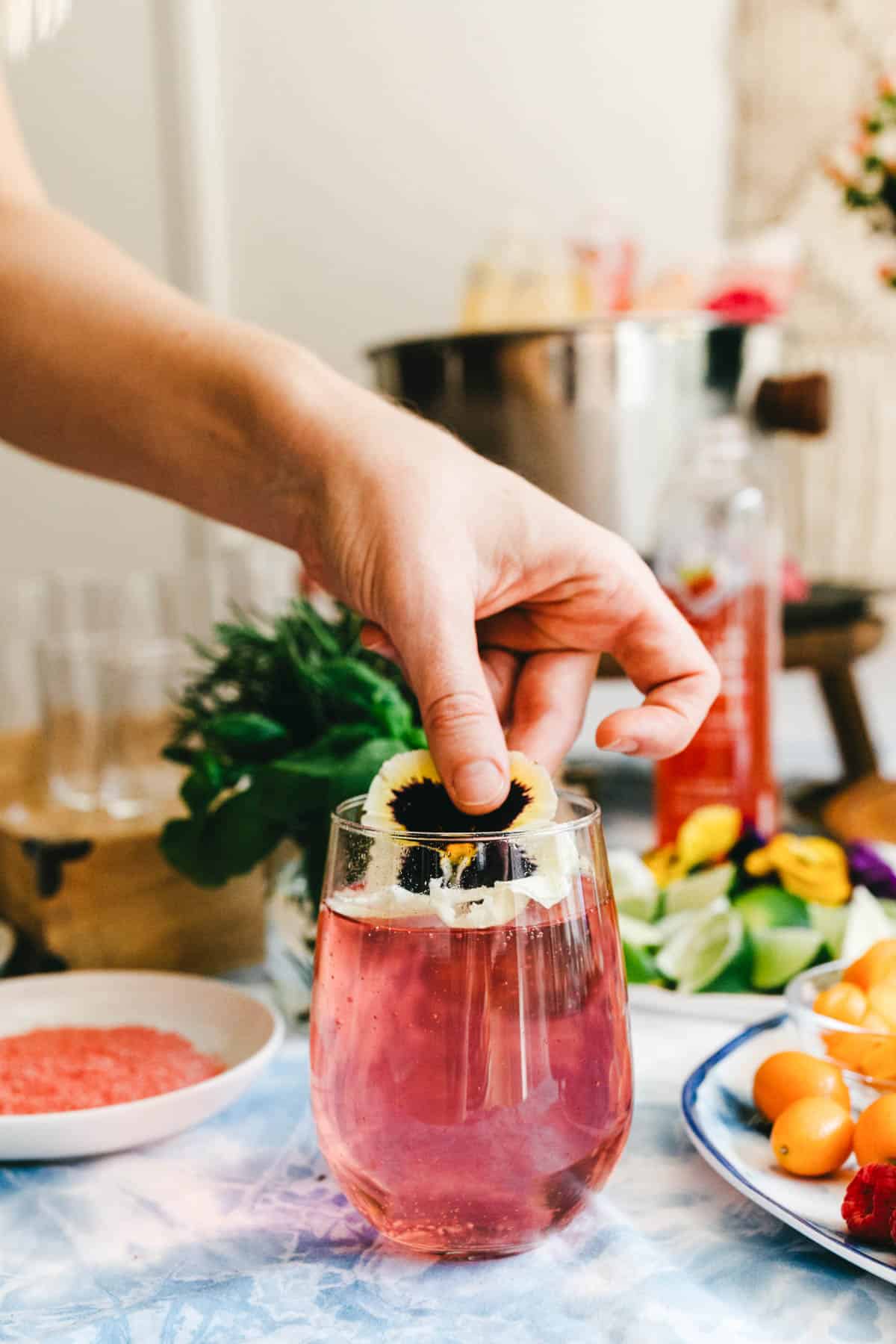 Woman adding an edible flower garnish to a pink cocktail on a party table. 
