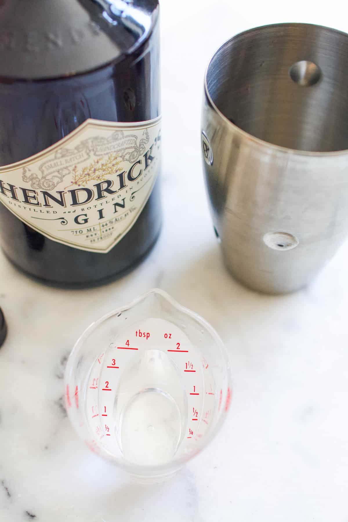 A measuring cup of gin next to a bottle of Hendrix gin and a cocktail shaker.