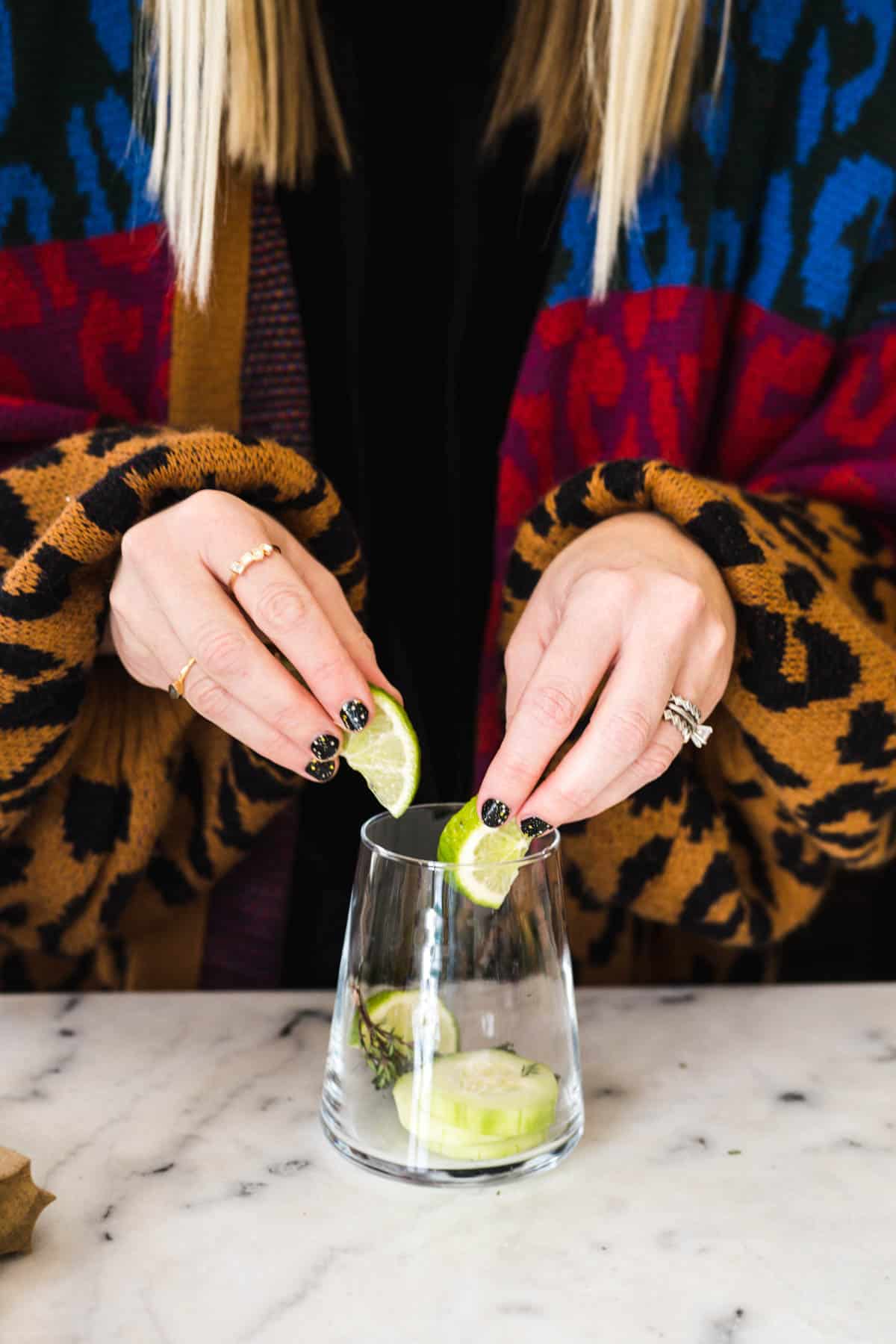 Woman putting lime slices, cucumber and fresh thyme into a cocktail glass.