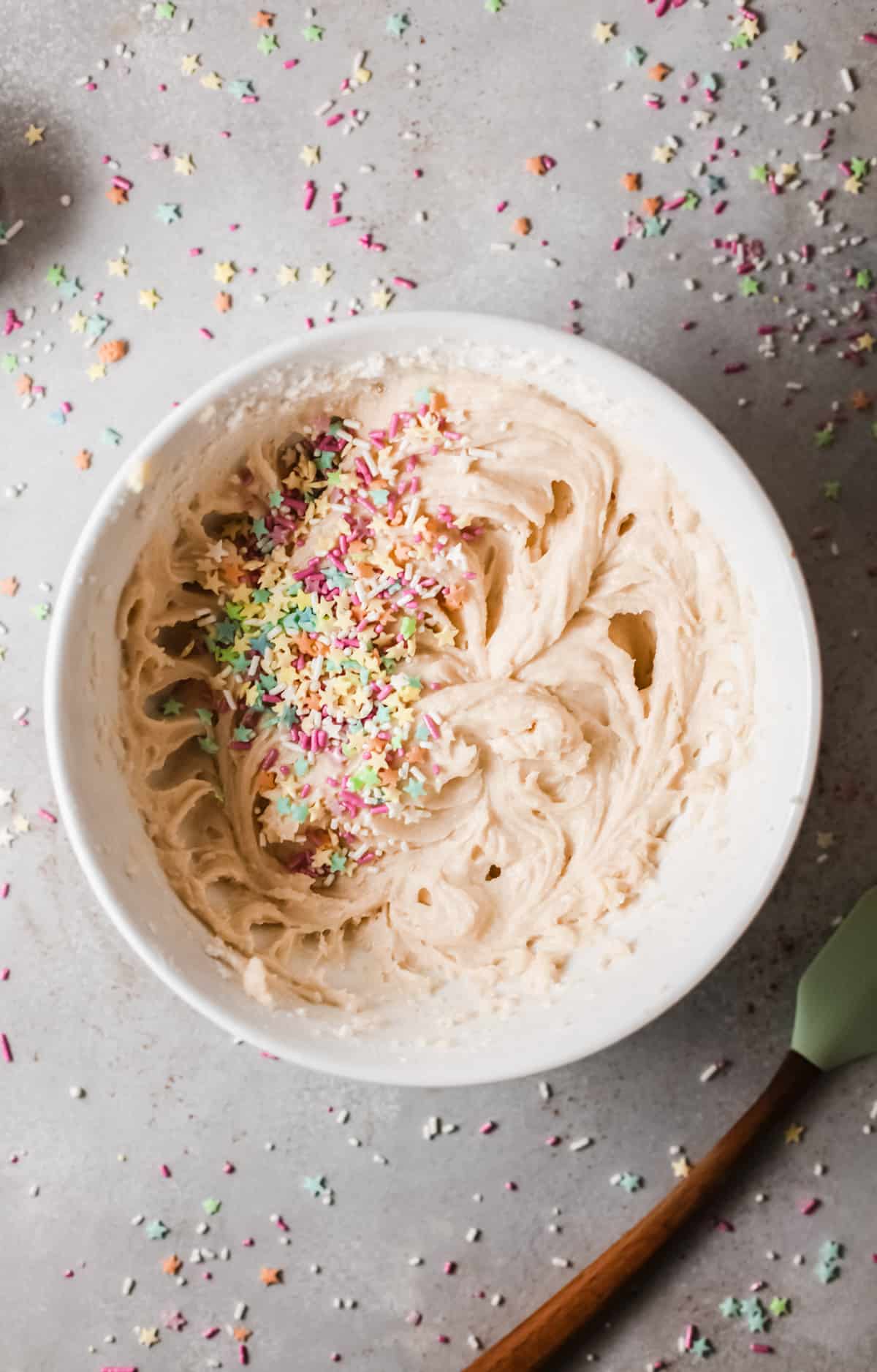 A bowl of vanilla cake mix topped with sprinkles next to a spatula on a gray countertop.