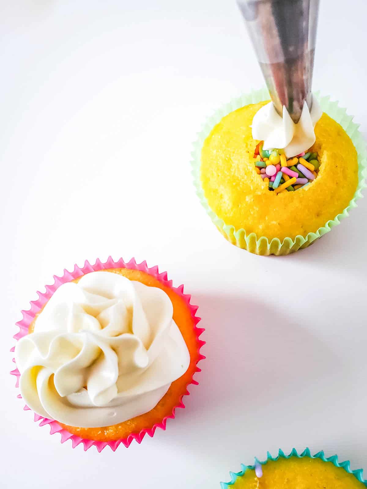 Cupcakes on a table with one being frosted to cover the hidden Easter sprinkles inside.