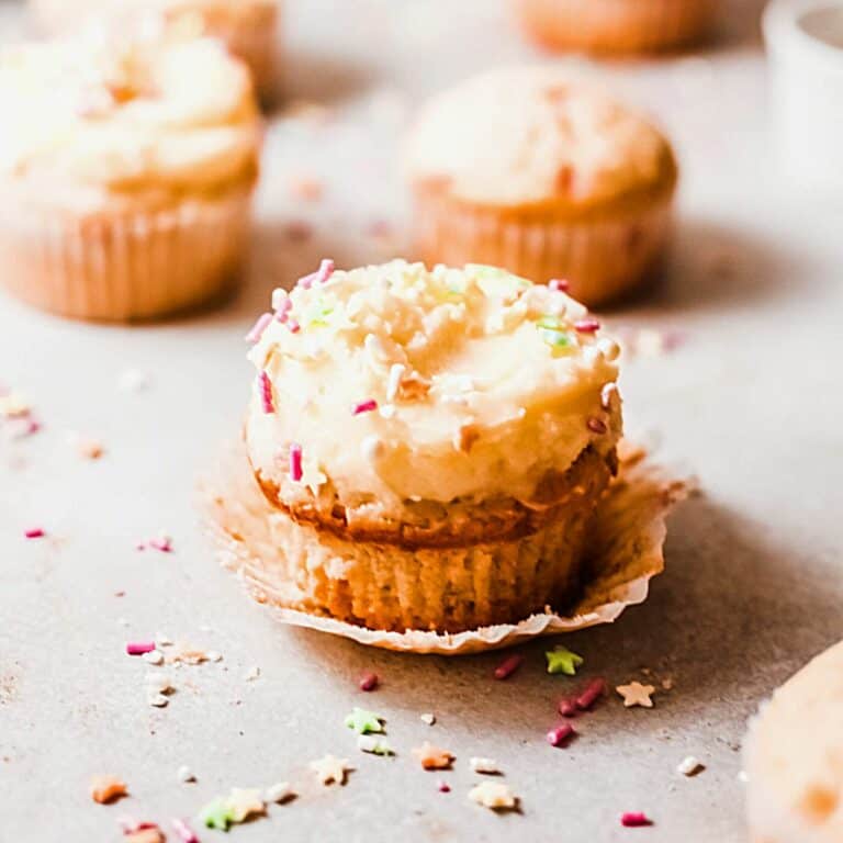 Sprinkle topped cupcakes in a paper wrapper sitting on a table with more cupcakes and sprinkles around it.