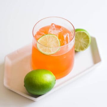 An Aperol summer cocktail in a glass on top of a white plate surrounded by fresh limes and topped with a lime slice.