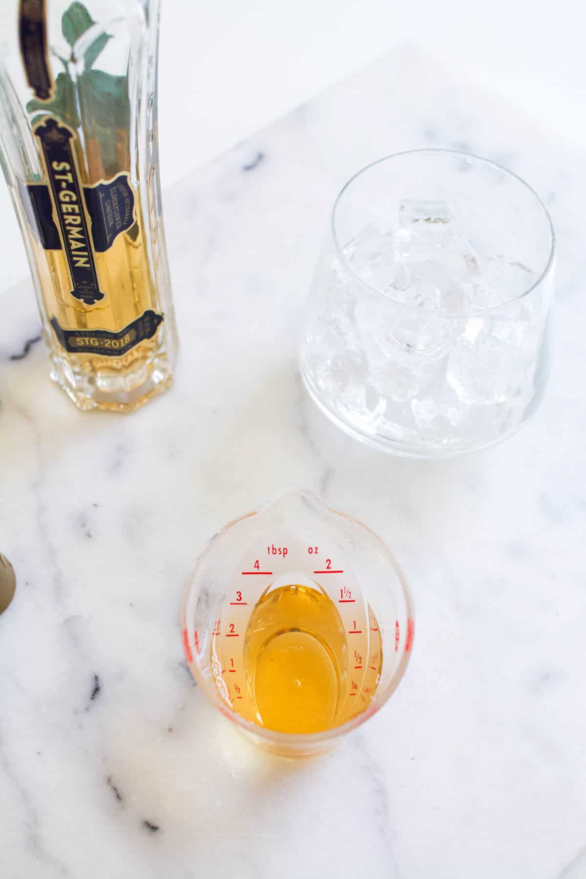 Bottle of St Germain, measuring cup of elderflower liqueur and a glass of ice on a countertop.