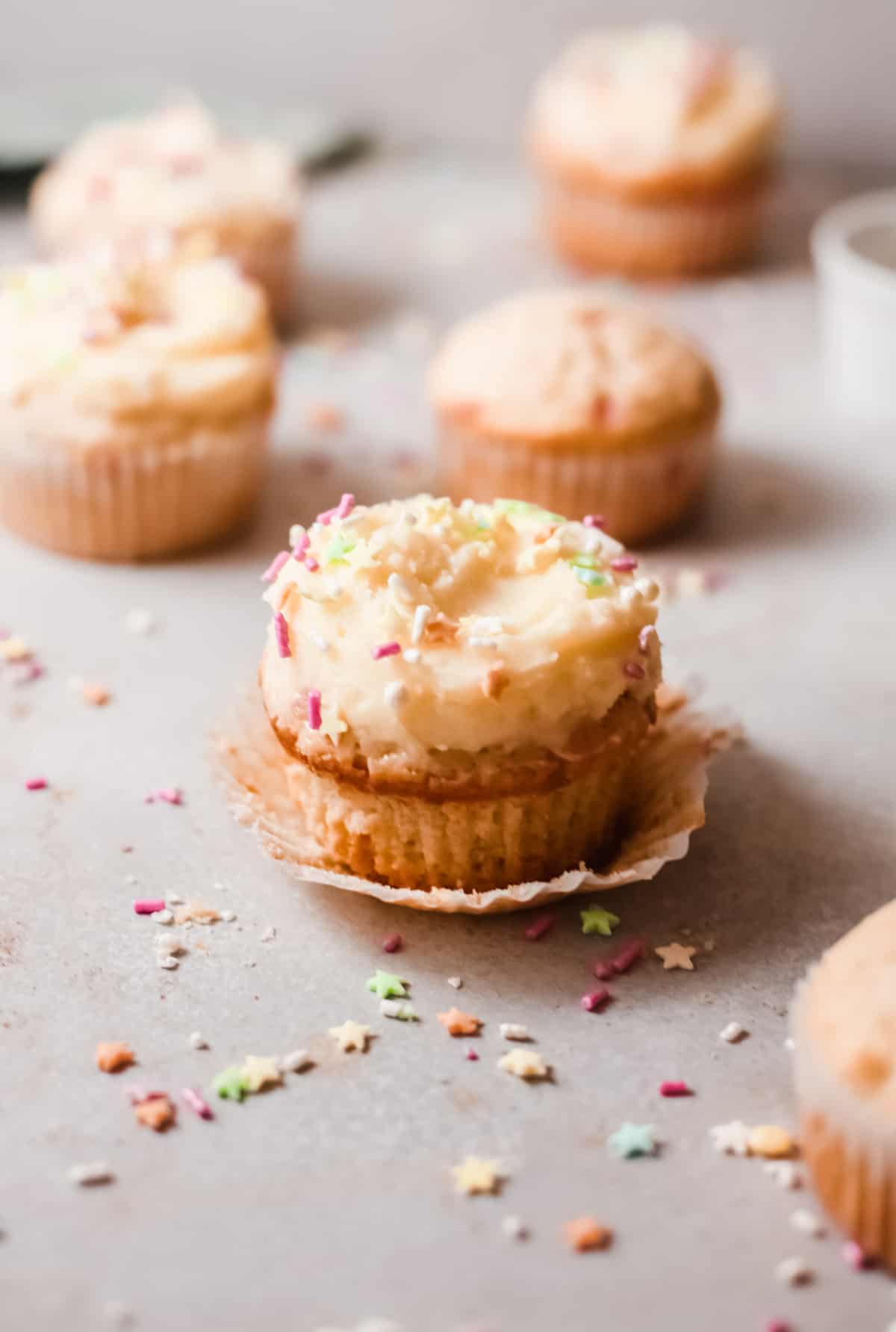 A birthday cupcake sitting in a torn off paper cupcake liner on top of a gray countertop and surrounded by more cupcakes and sprinkles.