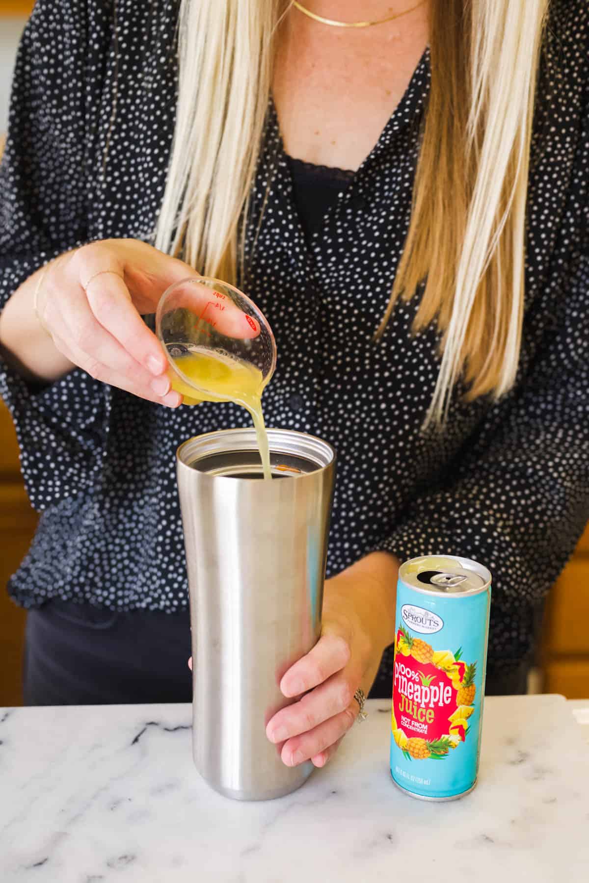 Woman pouring pineapple juice into a cocktail shaker from a small measuring cup.