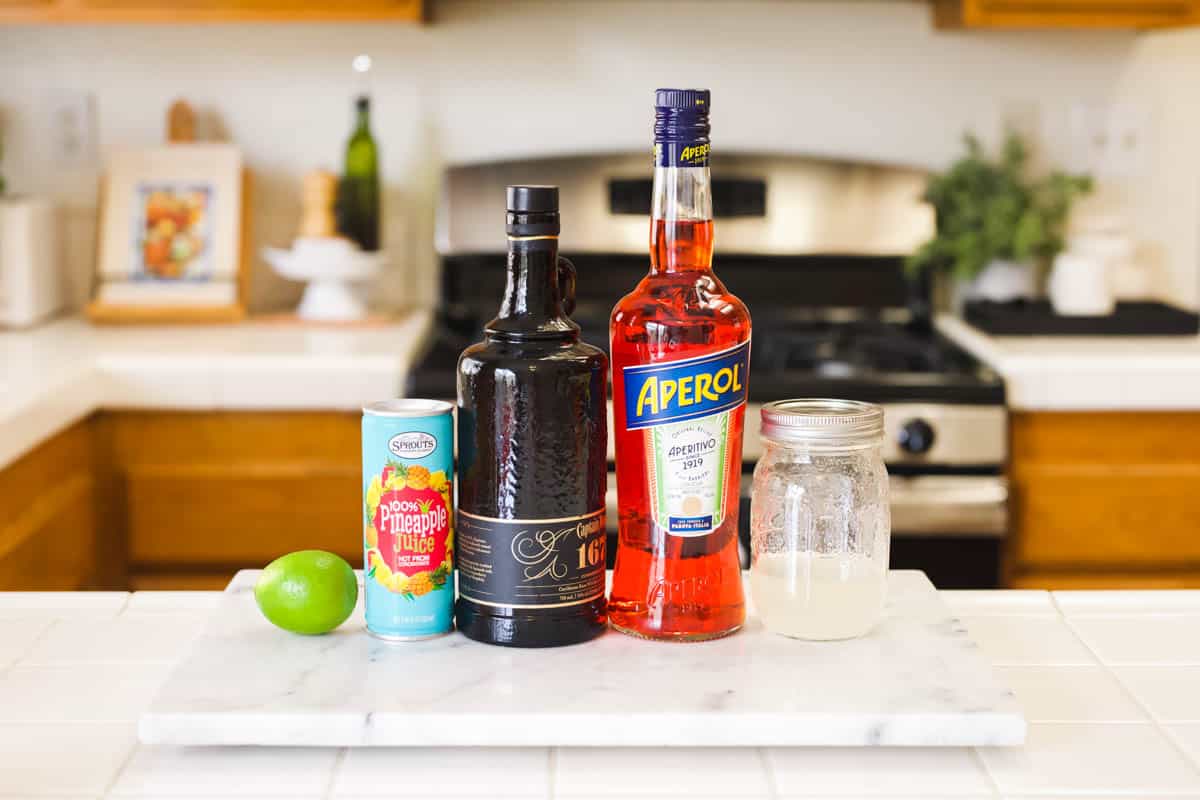 Ingredients to make a tropical spiced rum Aperol cocktail - lime, pineapple juice, spiced rum, Aperol liqueur, and simple syrup on a white marble counter top.