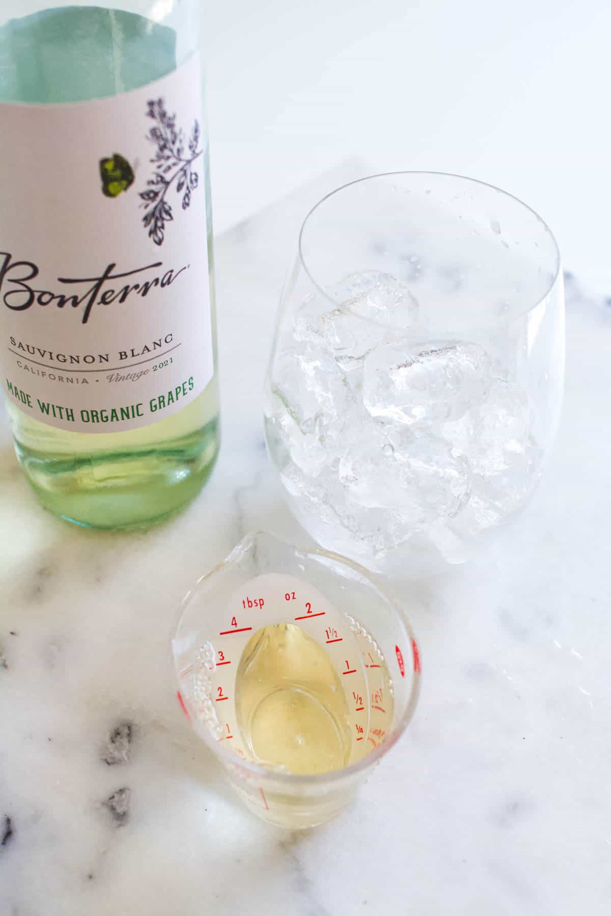 White wine in a clear measuring cup in front of a glass filled with ice and a bottle of white wine.