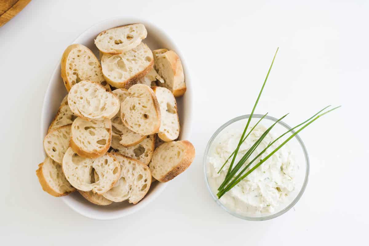 A bowl of crostini bread next to a small glass bowl of burrata dip on a white countertop.