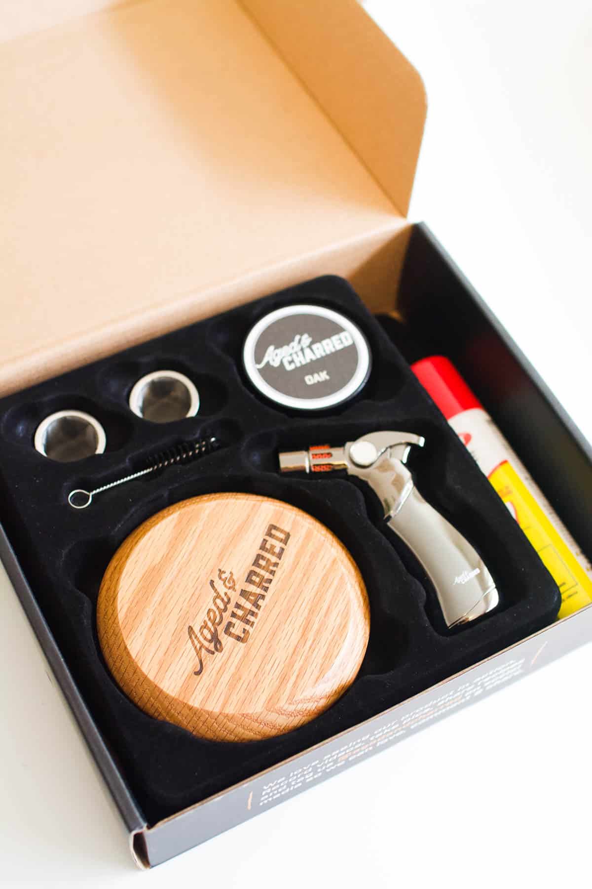 The inside of the Aged and Charred cocktail smoker kit box holding butane, a torch, a cocktail smoker, and wood chips.