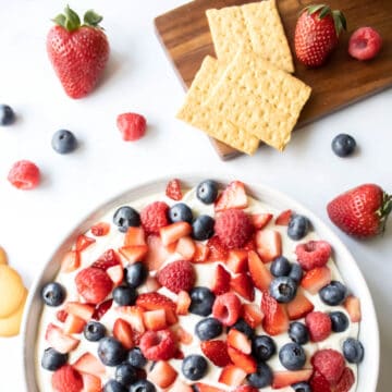A bowl of blueberries, raspberries and strawberries on a cheesecake dip next to fruit and graham crackers.