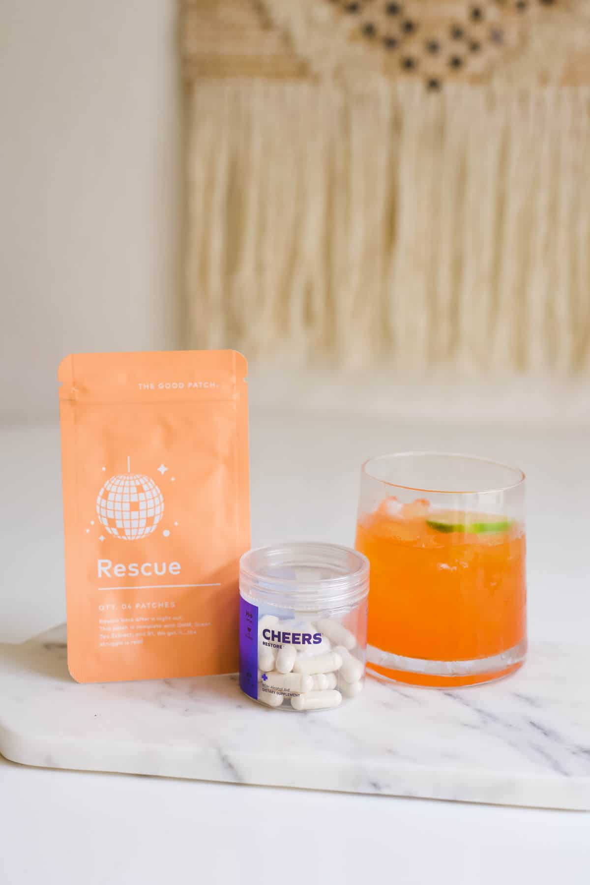 A highball glass filled with a cocktail on a marble cutting board next to a bottle of hangover supplements and a Rescue Patch.