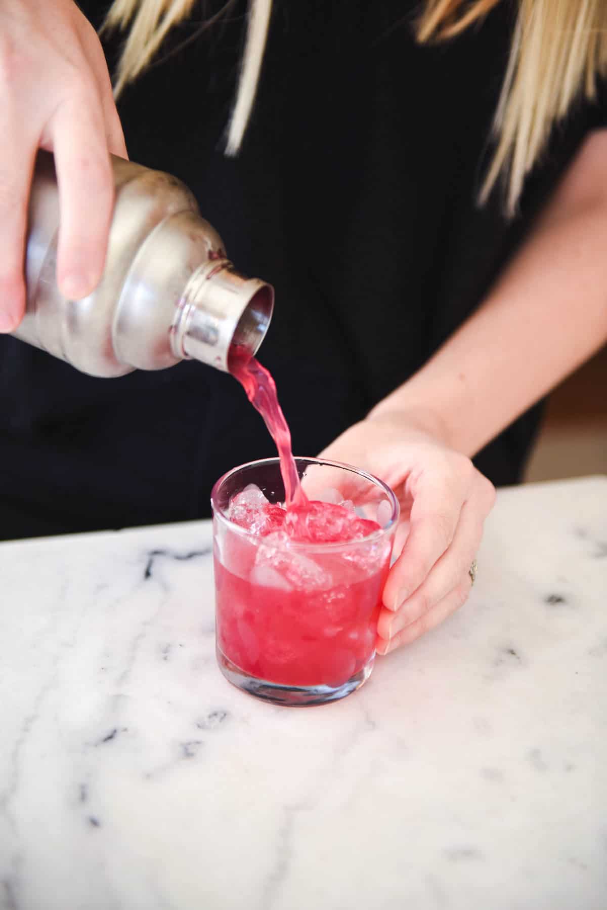 A woman pouring a pink cocktail into a highball glass.