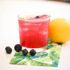 A square image of a bright pink blueberry lemonade vodka cocktail surrounded by fresh fruit.
