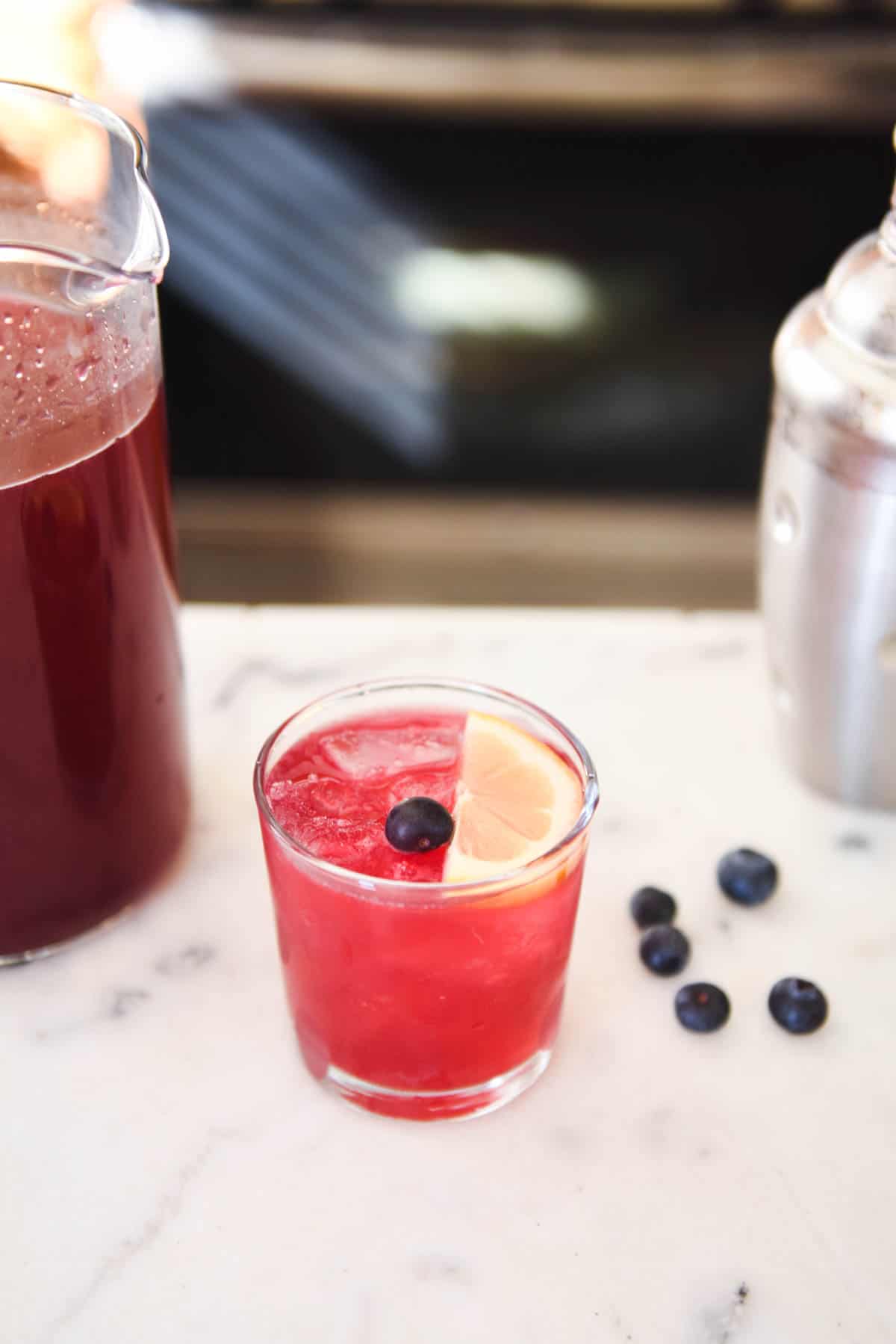 A blueberry lemonade vodka cocktail between a cocktail shaker and a pitcher of blueberry lemonade.