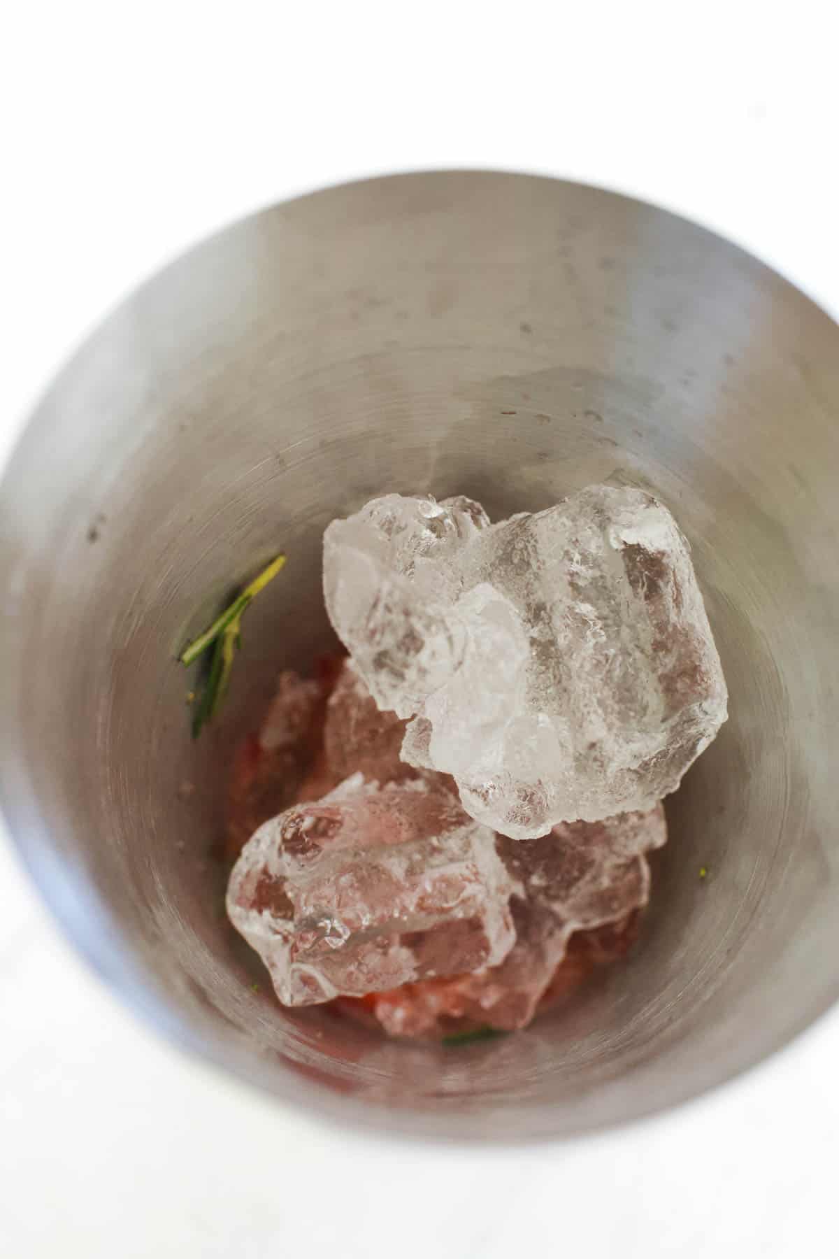 Astainless steel cocktail shaker filled with strawberries, rosemary and ice.