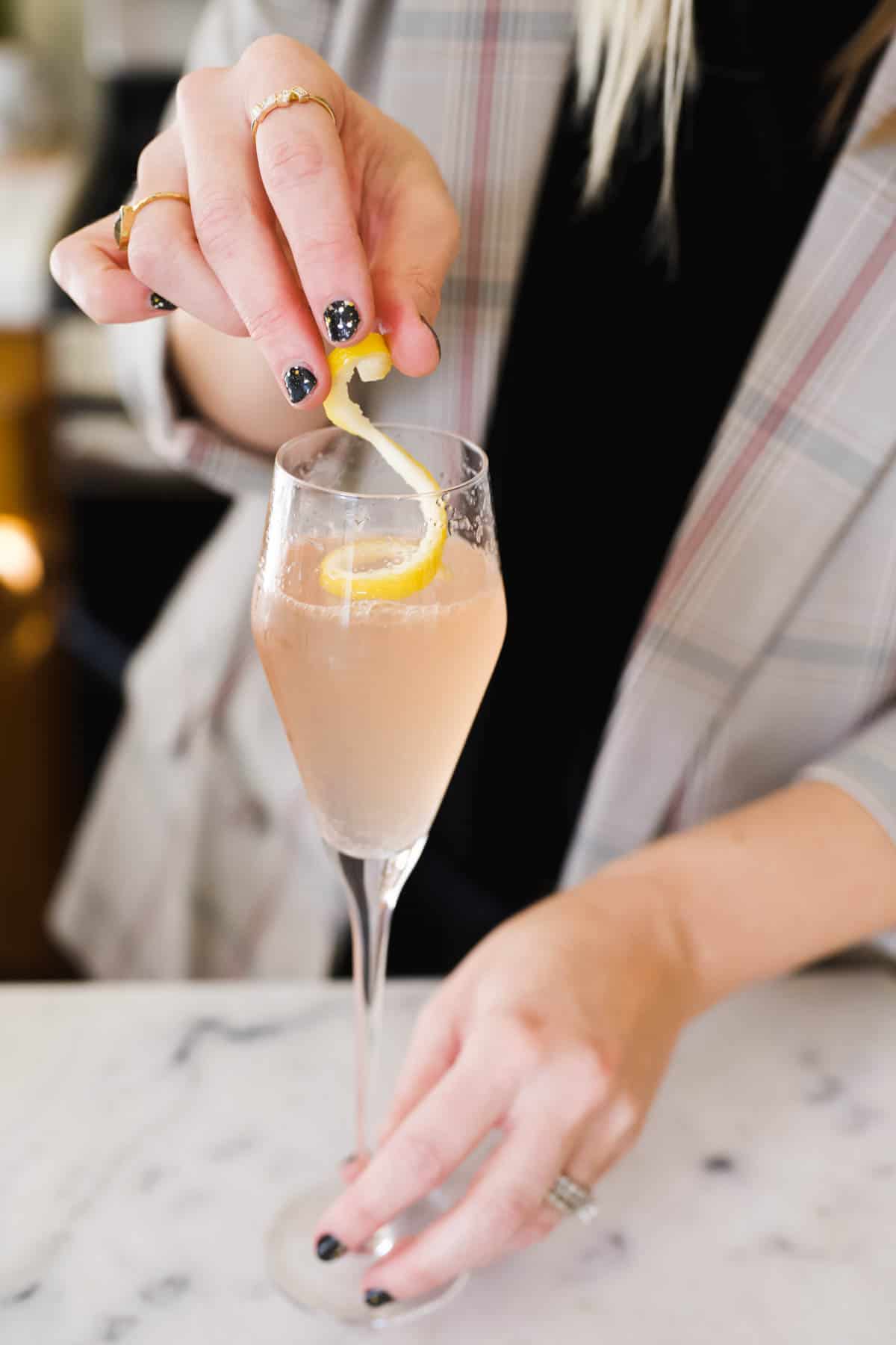Woman adding a lemon twist to a pink champagne cocktail in a champagne flute.