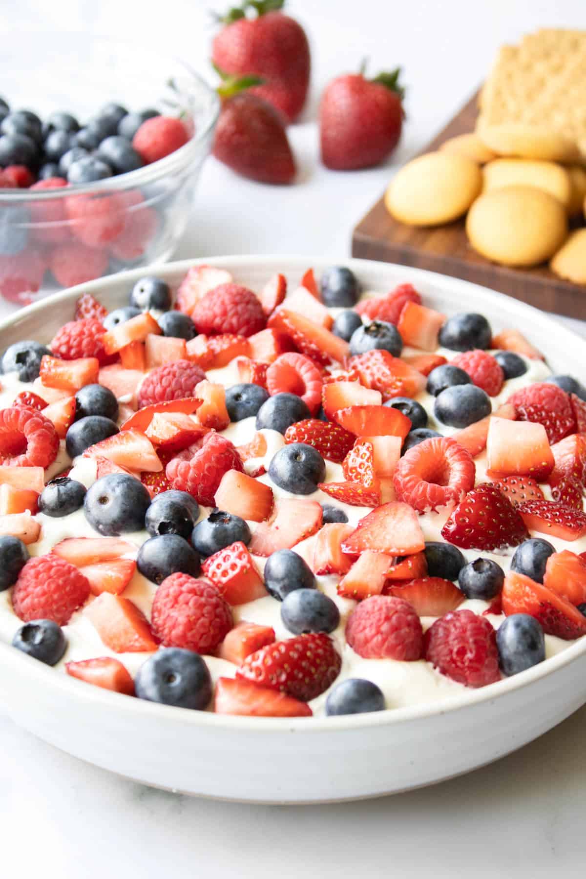 A bowl holding cheesecake dip topped with red and blue berries and cookies to dip in it in the background.