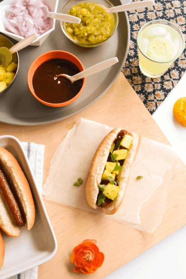 A hot dog topped with BBQ sauce and pineapple on a table next to condiments for a party.