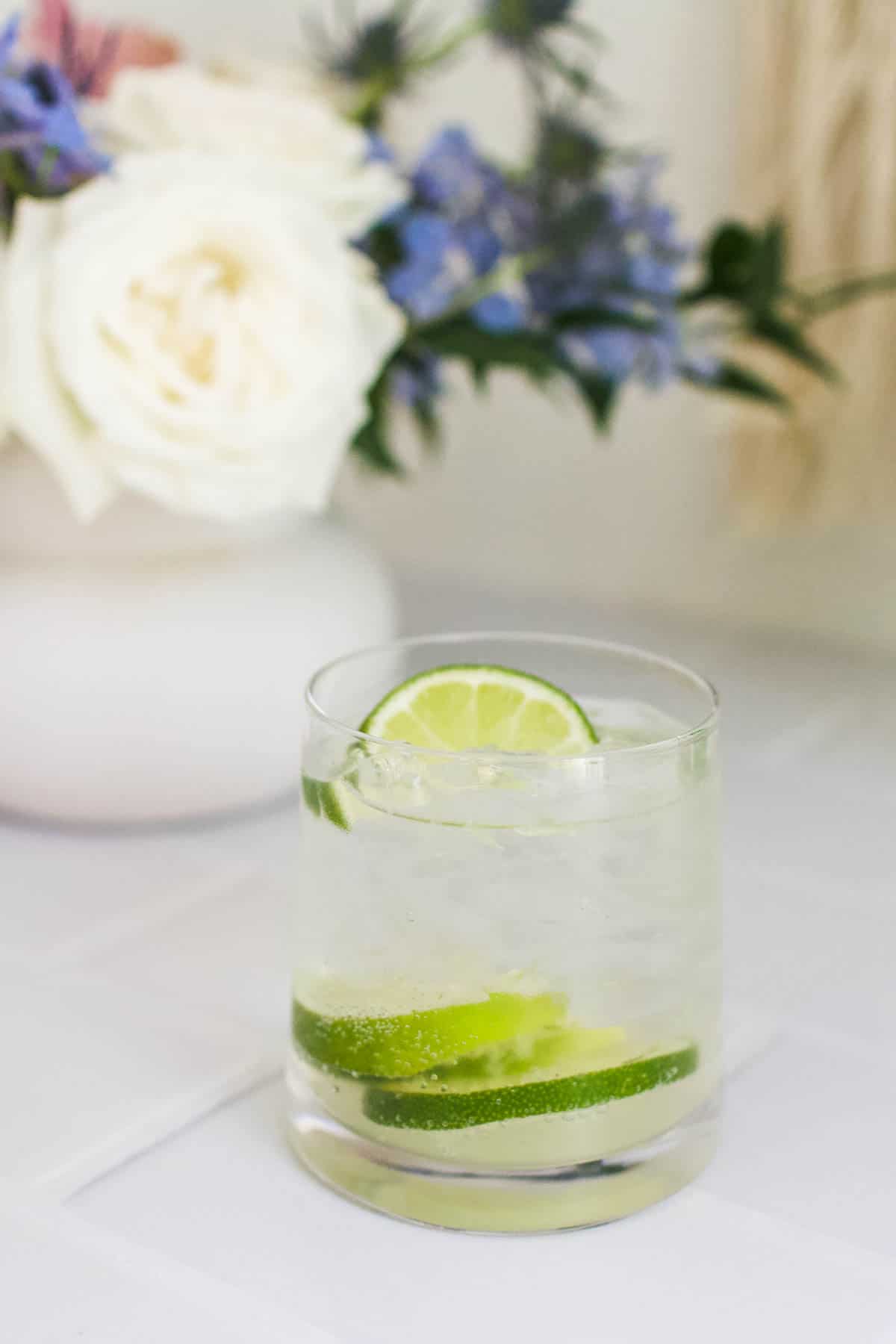 Gin and tonic garnished with lime with a vase of flowers in the background.