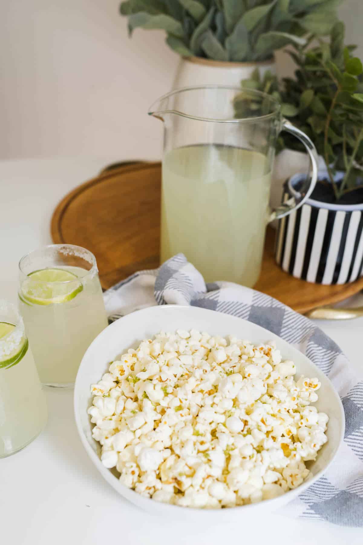 A bowl of popcorn on a table with a pitcher of margaritas and two glasses of margaritas.