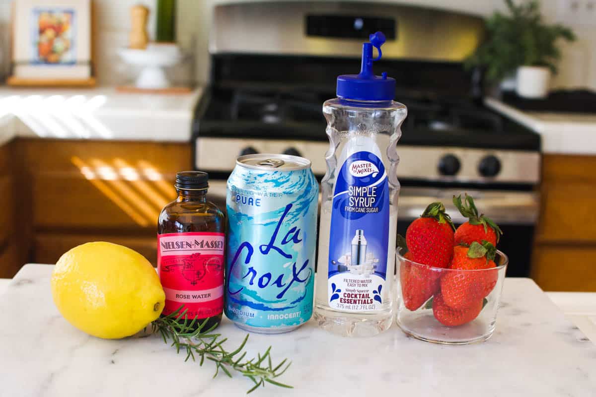 Ingredients to make a strawberry mocktail including strawberries, rosemary, fresh lemon, bitters, sparkling water, and simple syrup.