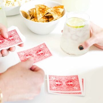A person dealing cards for two people while enjoying snacks and margaritas.