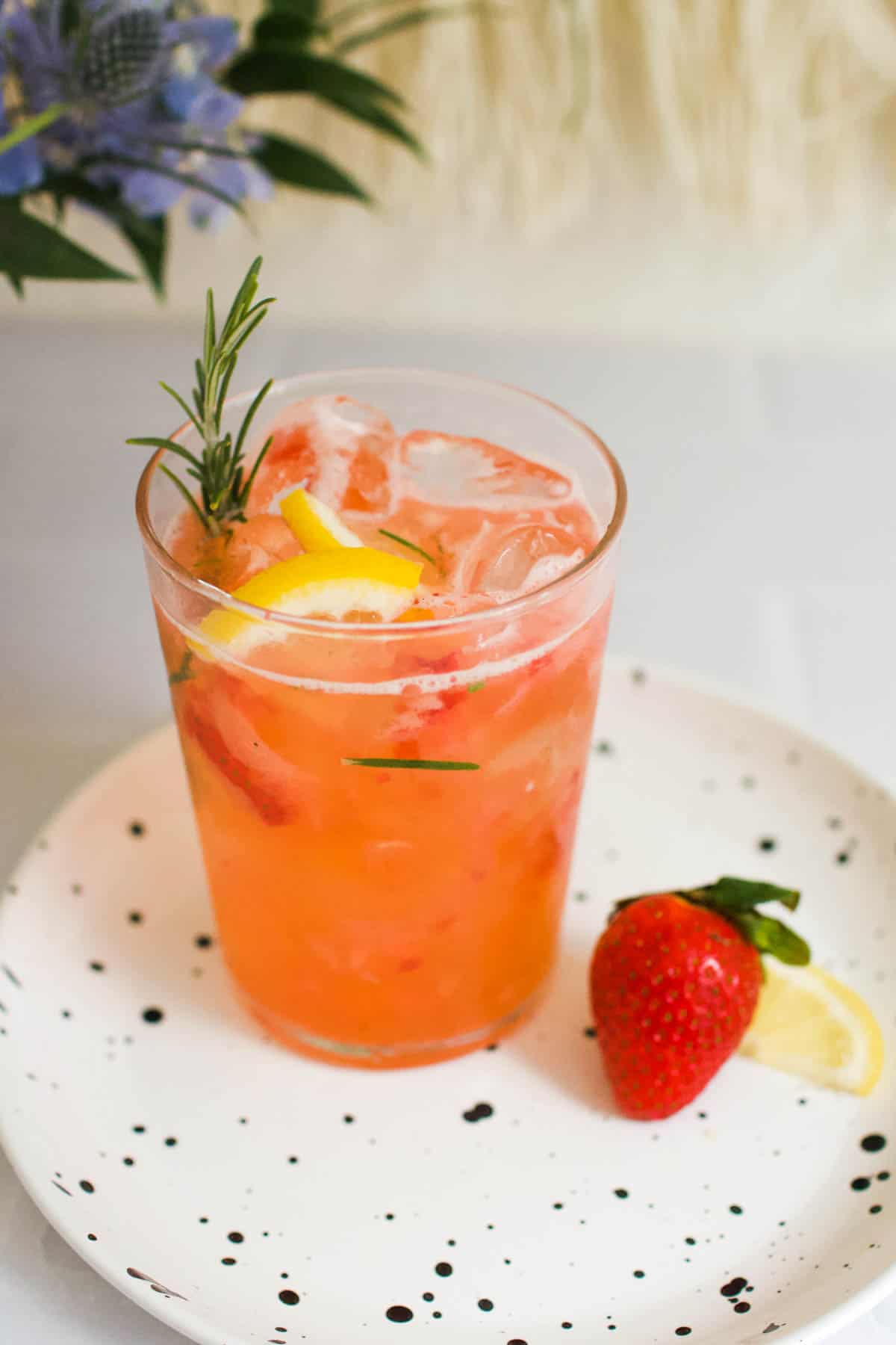 A mocktail with strawberries and fresh rosemary on a plate next to a fresh strawberry.
