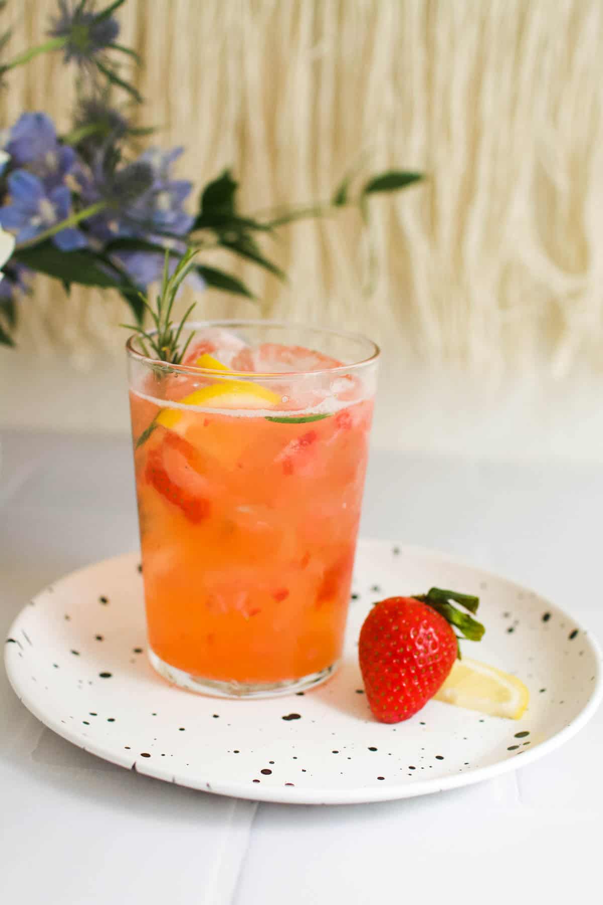 A strawberry mocktail in a glass topped with fresh rosemary and lemon slices next to fresh strawberries on a plate.