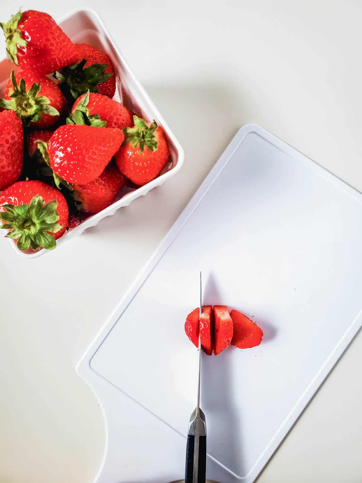 A white cutting board with a strawberry being sliced next to a bowl of more strawberries.